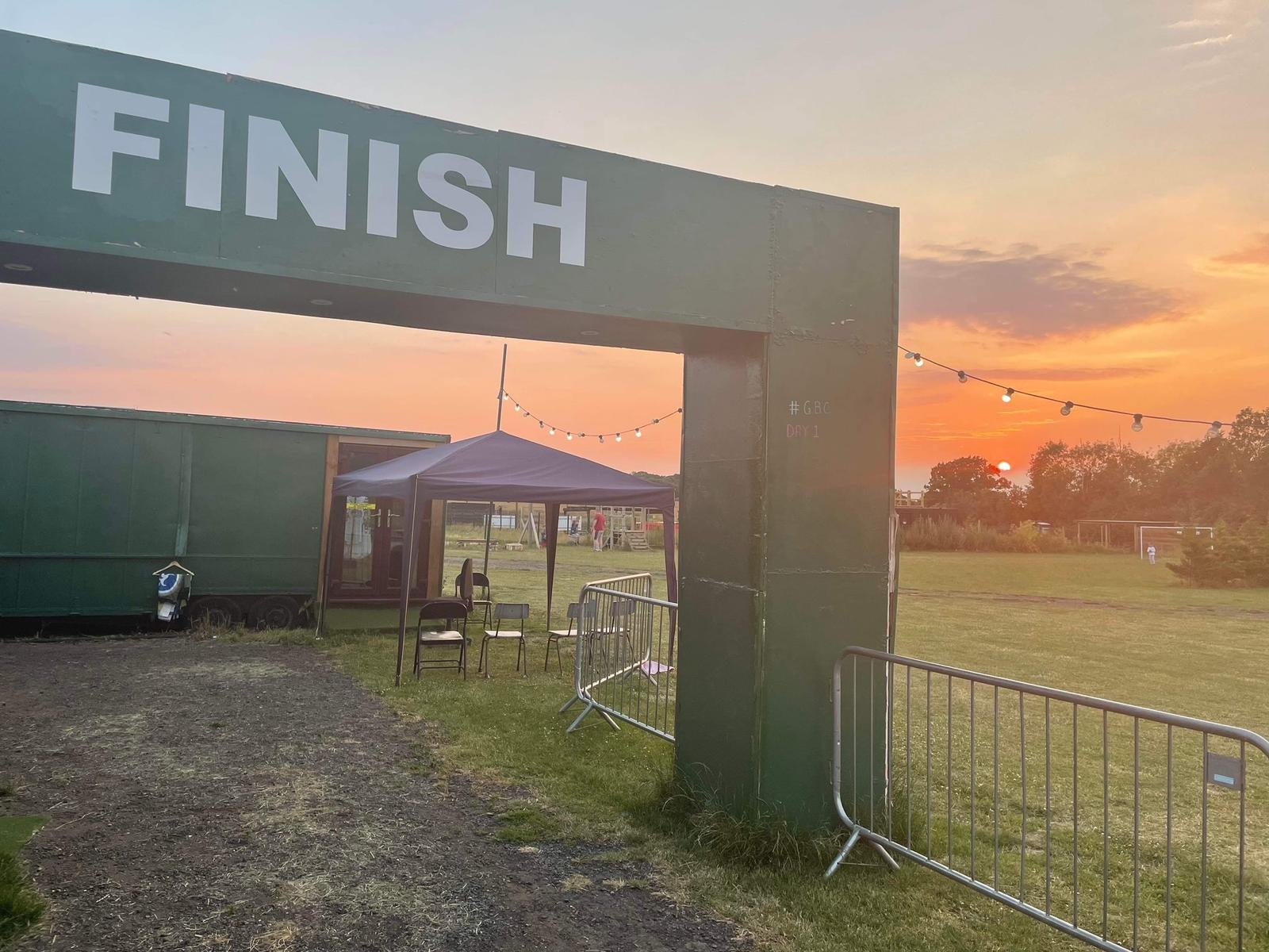 A green metal finish line structure stands on a grassy field at sunset. There are chairs and tables under a nearby canopy, and string lights hang above. Metal barriers line the path under the finish structure, and the sky glows with purple and orange hues.