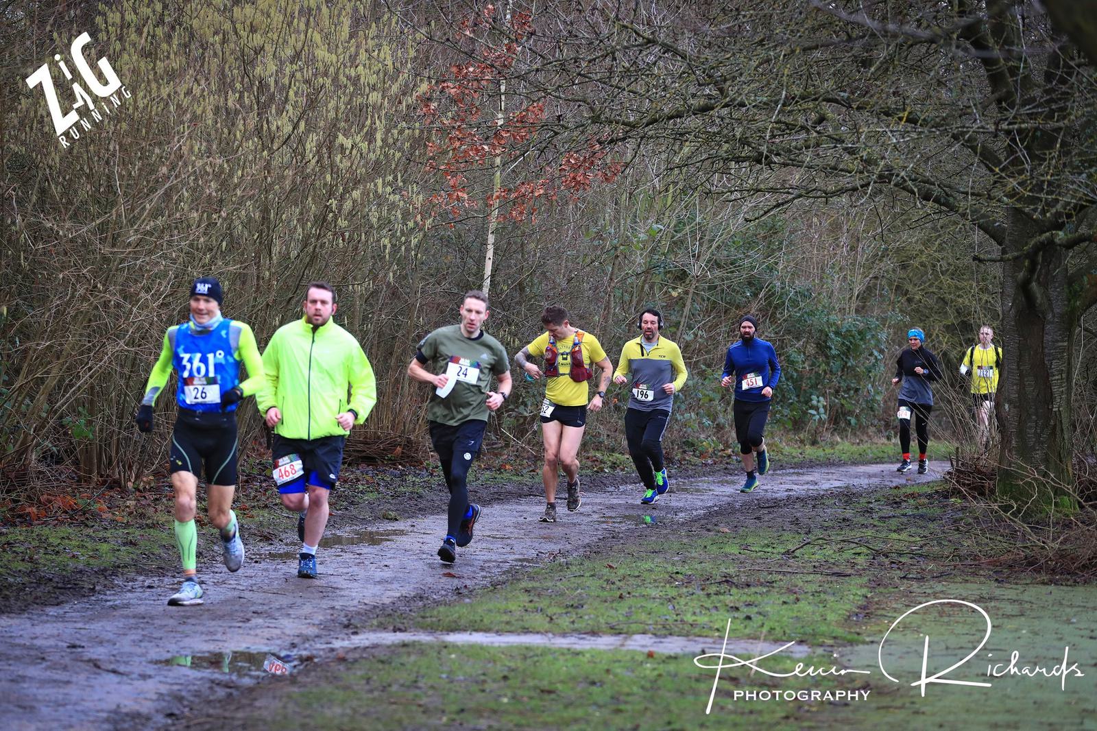A group of runners participating in a trail race on a muddy path surrounded by trees. They wear various athletic gear, including bright shirts and shorts. The scene appears lively and energetic amidst the natural setting.