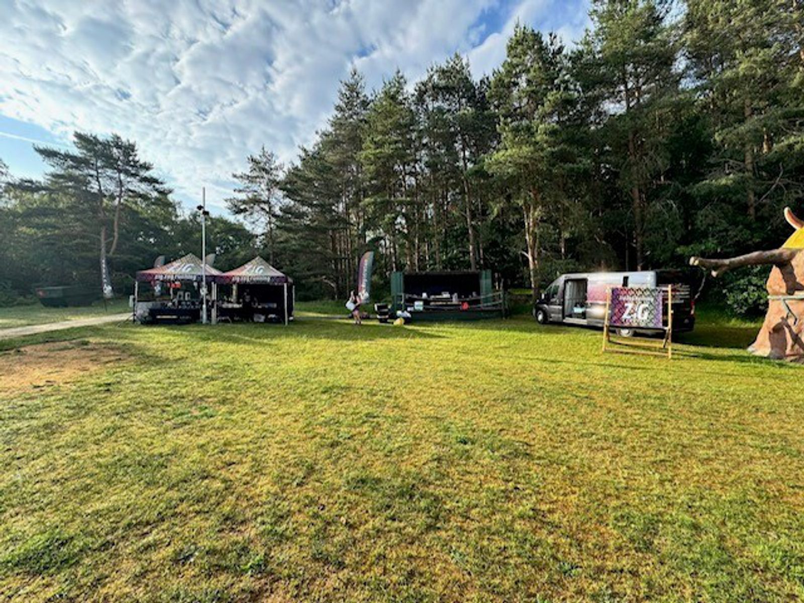 A grassy open field with food trucks, tents, and a van set up near the edge of a forest under a partly cloudy sky. A large wooden sculpture is visible on the right side of the image.