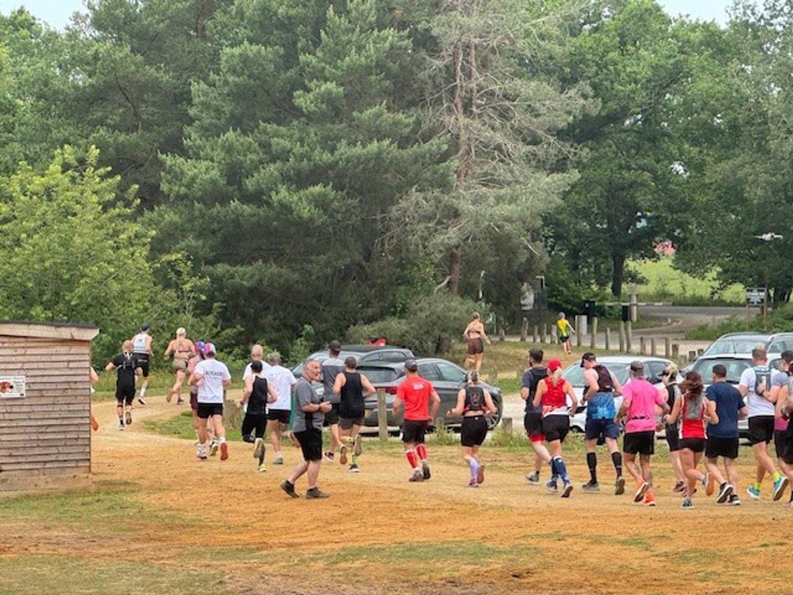 A group of runners in athletic gear participate in an outdoor race or fun run on a grassy path with trees and parked cars in the background.