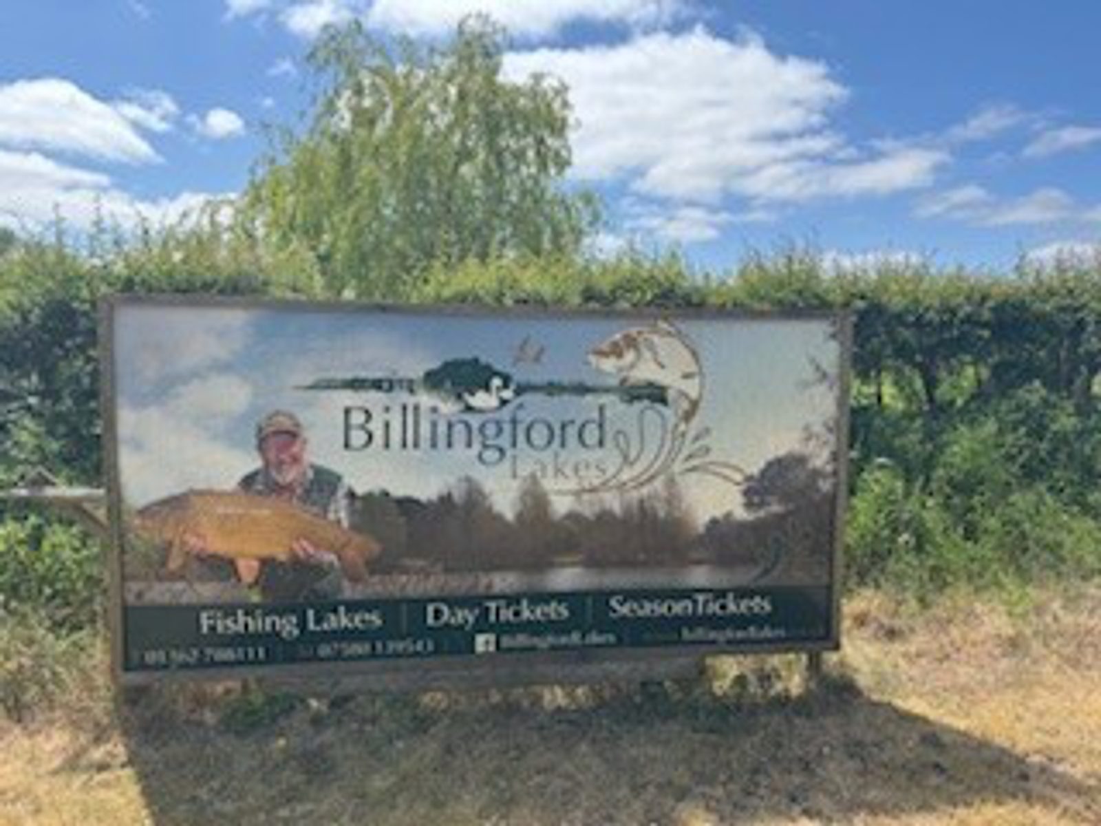 A sign for Billingford Lakes shows a man holding a large fish, with trees and a blue sky in the background. The sign advertises fishing lakes, day tickets, and season tickets.