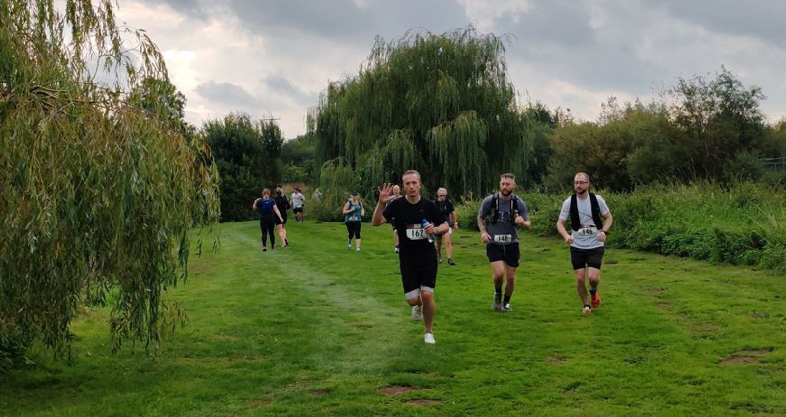 A group of runners participates in a race on a grassy path surrounded by trees. The lead runner is waving and smiling at the camera, while others follow closely behind under a cloudy sky.