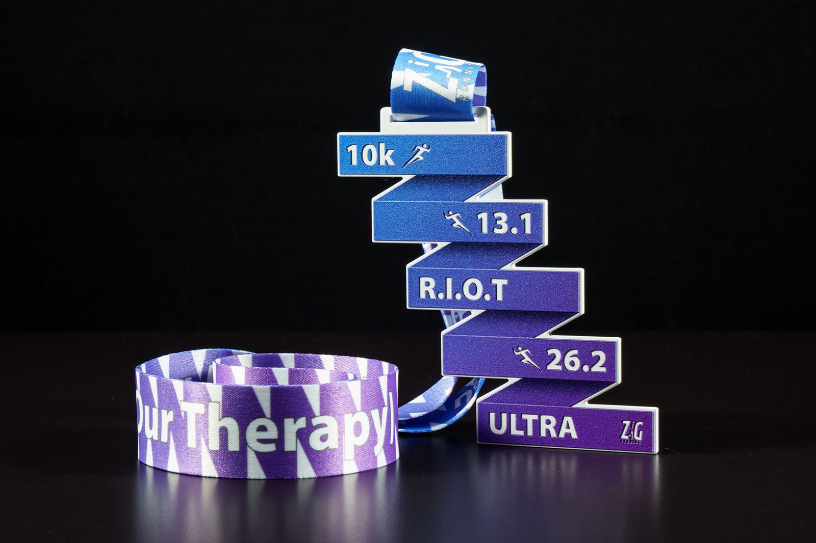A vivid, colorful race medal displaying distance markers including 10k, 13.1, R.I.O.T, and 26.2 on a staircase-shaped design. The connected purple ribbon has a white zigzag pattern and partially visible text, "YourTherapy," all set against a black background.