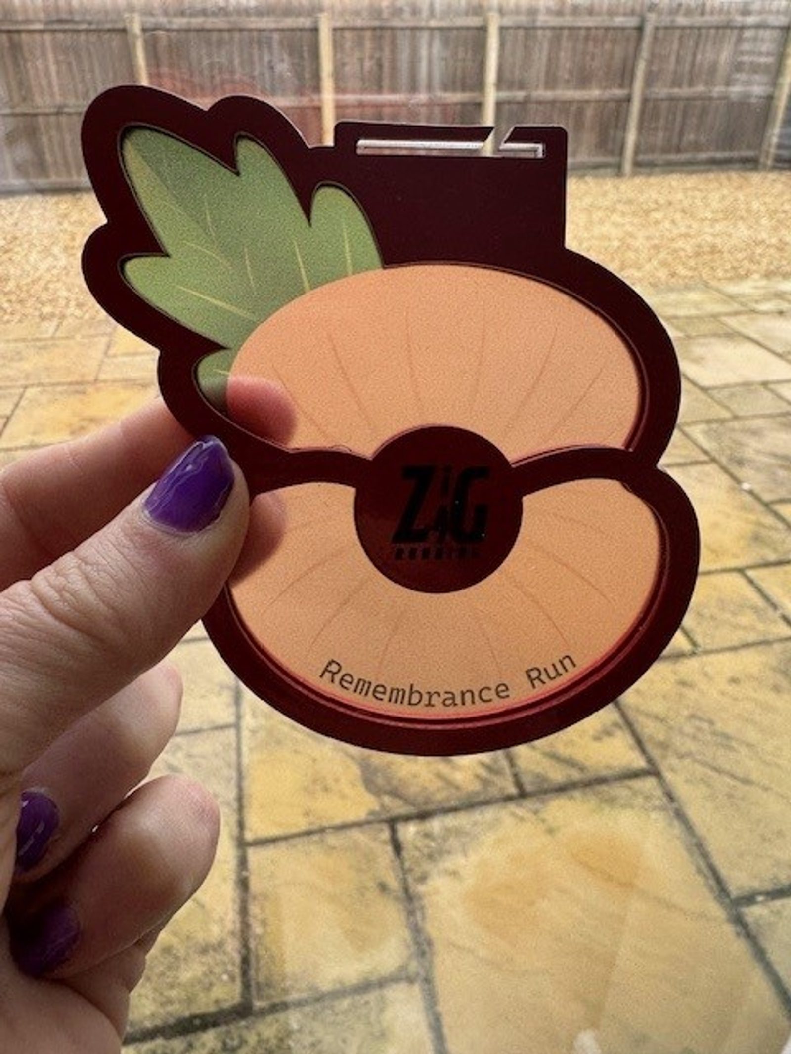 A hand with purple nail polish holds a poppy-shaped medal with a green leaf. The medal reads "Remembrance Run" and "Zig." A stone patio and fenced yard are visible in the background.