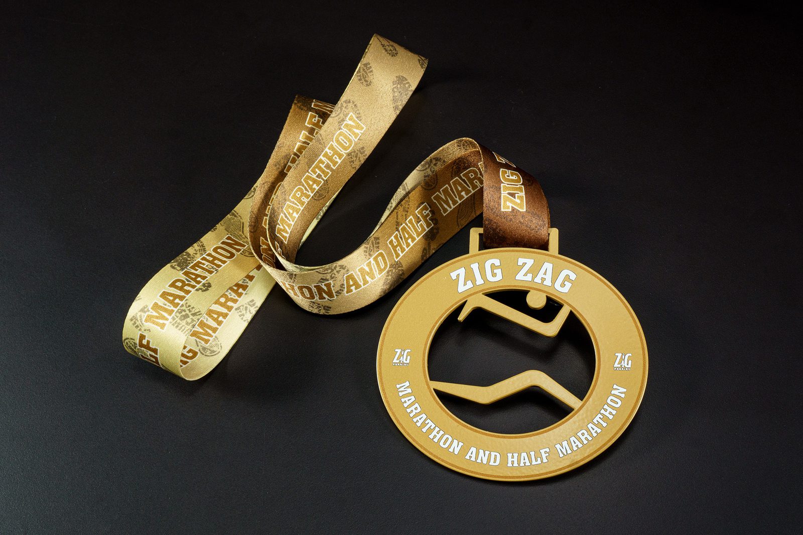 A gold medal hangs from a brown ribbon with "Marathon and Half Marathon" text repeatedly printed on it. The medal has "Zig Zag" at the top and "Marathon and Half Marathon" at the bottom, with a zigzag line design in the center.