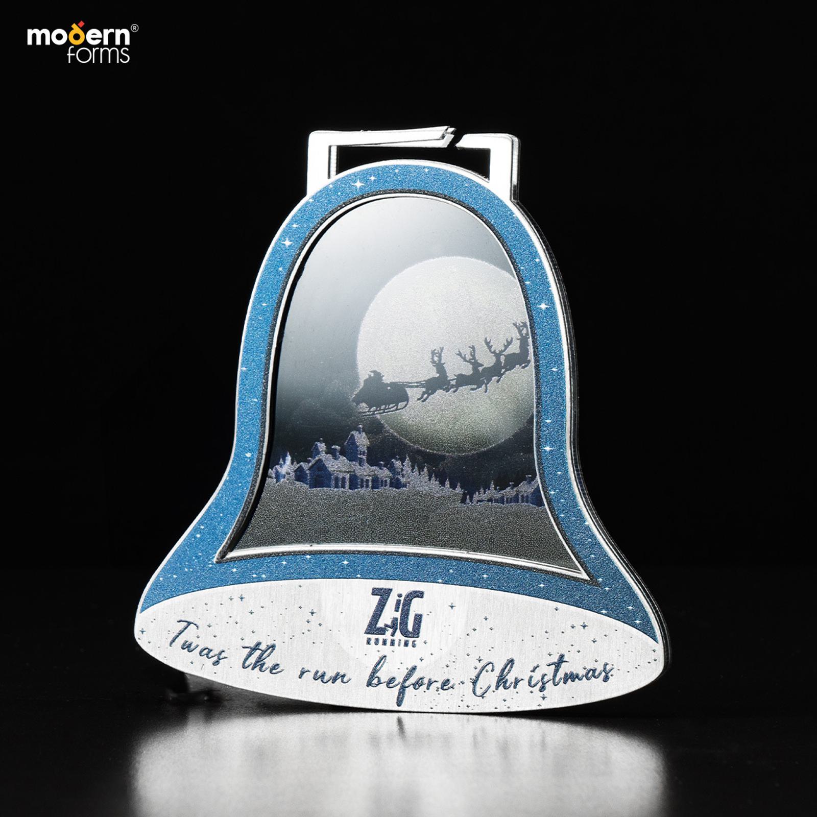 A bell-shaped Christmas ornament with a snowy cityscape and Santa's sleigh silhouetted against a large moon. The blue and silver ornament features the text "Zig" and "Twas the run before Christmas.