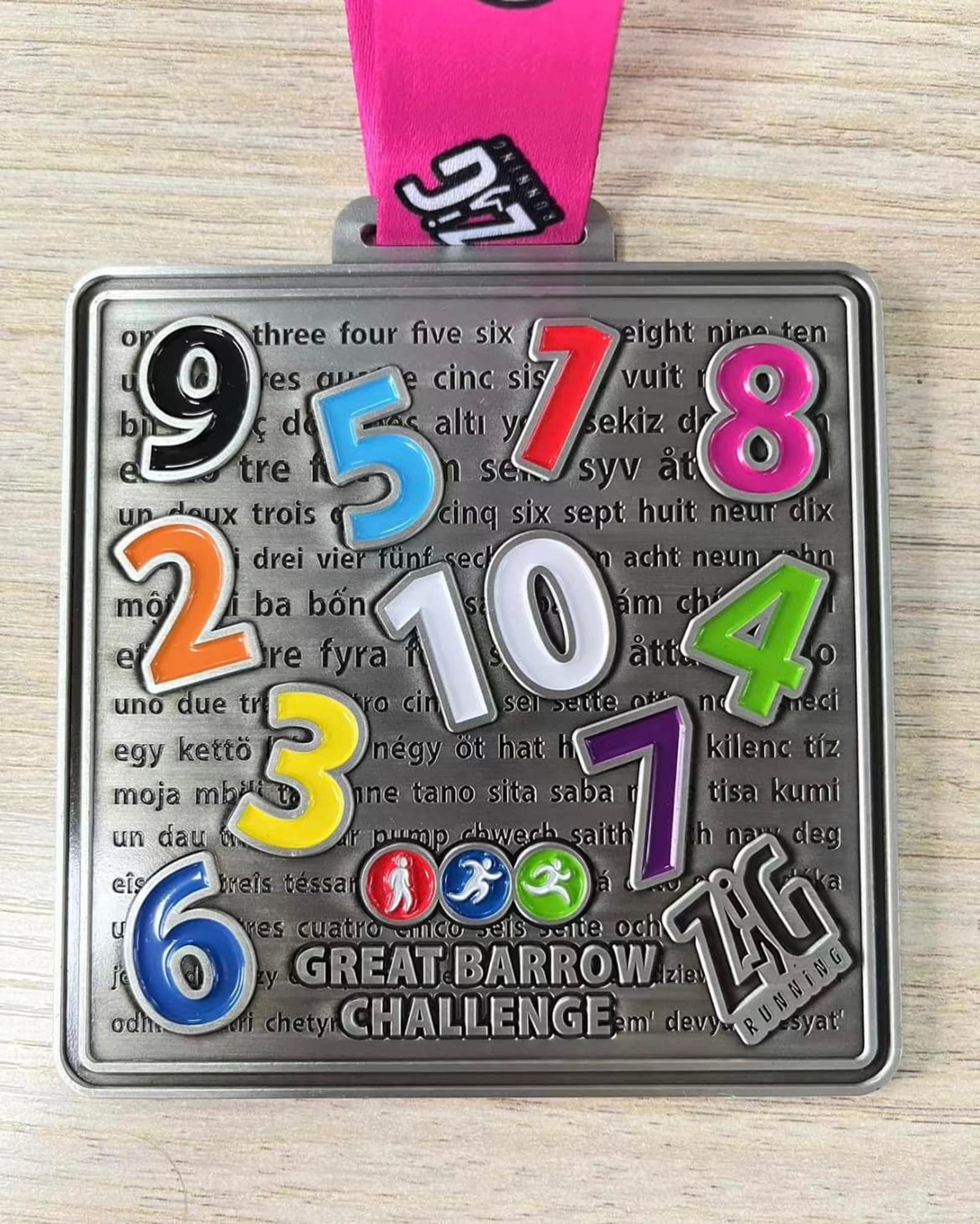 A rectangular medal with colorful numbers 1 to 10 in various fonts. The background has the same numbers written in different languages. At the bottom, text reads "Great Barrow Challenge" above logos of a runner, cyclist, and walker, and "TAG Running." It has a pink lanyard.
