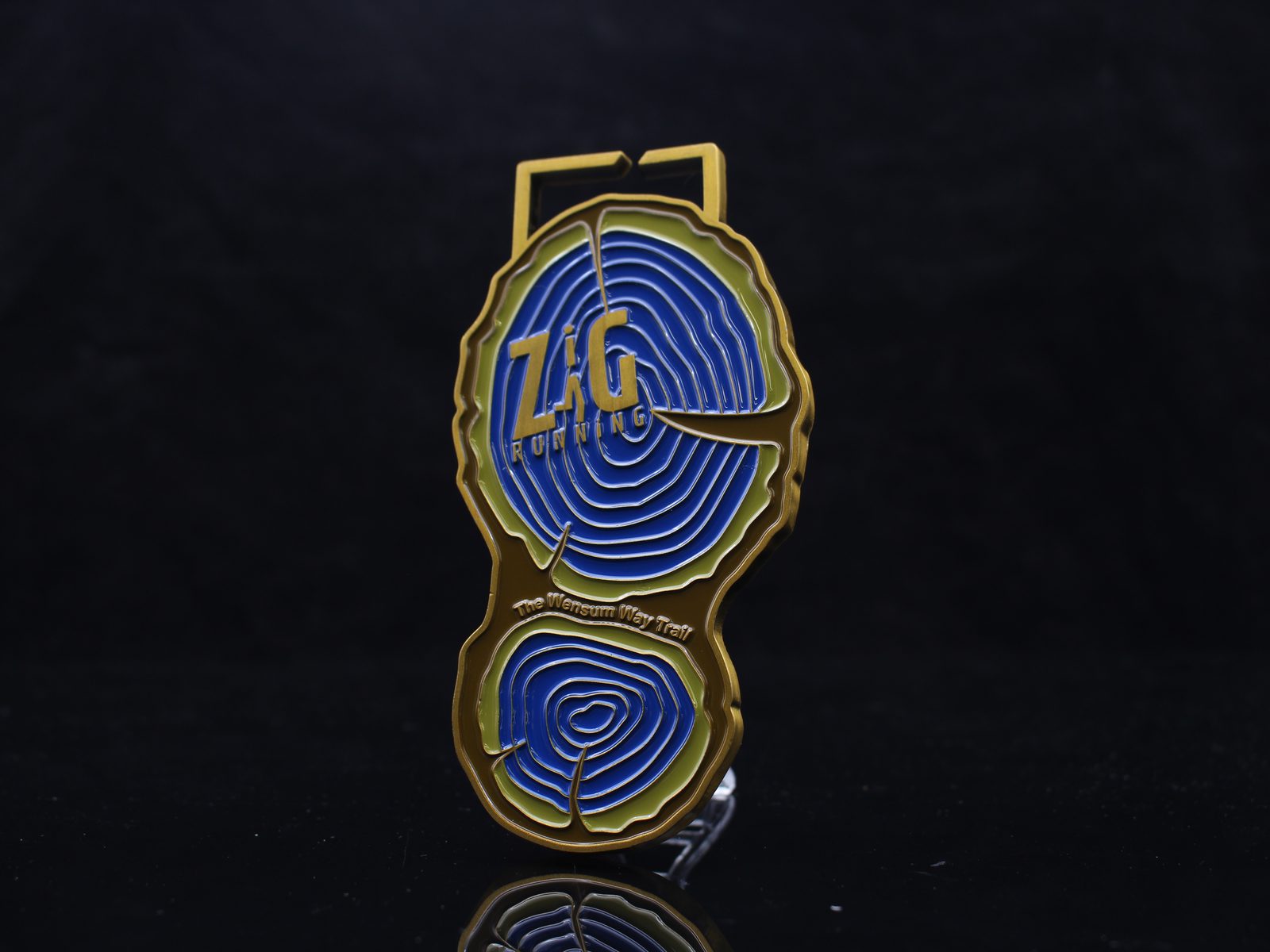 A gold and blue race medal shaped like tree rings, with the text "ZAG Running" in the center and "The Magnum Way Trail" at the bottom, set against a black background.