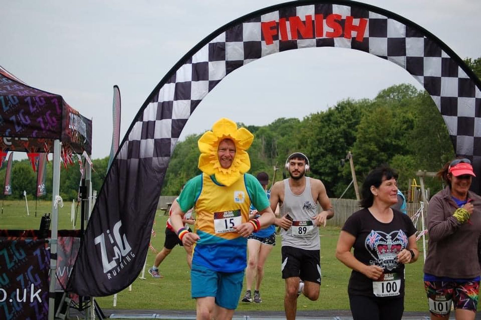 A runner dressed in a colorful costume resembling a flower crosses the finish line of a race. Other participants are also completing the race, with a checkered arch marking the finish. The event appears to be outdoors on a grassy area with trees in the background.