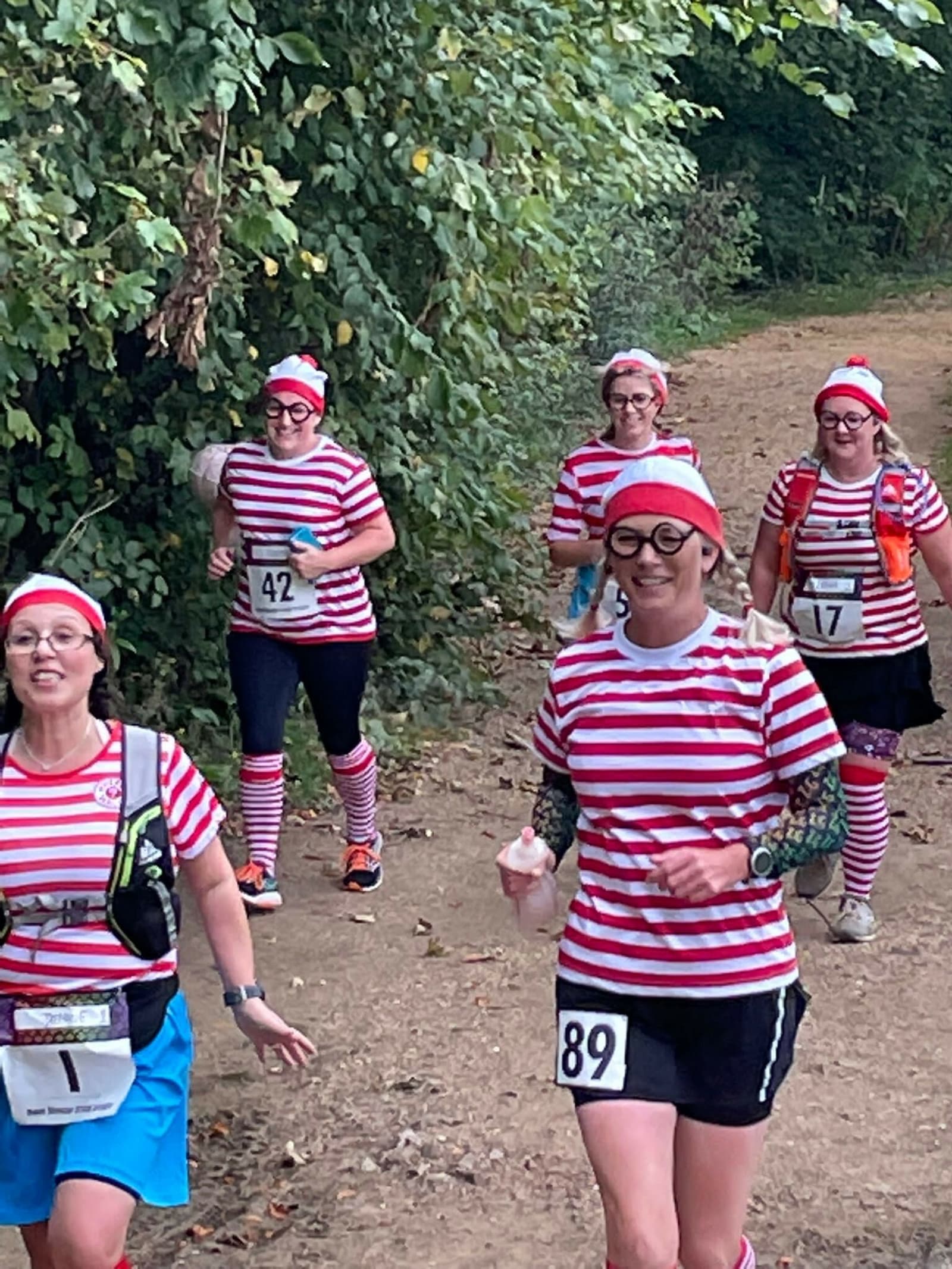A group of runners dressed in red and white striped shirts, red and white beanie hats, knee-high striped socks, and glasses run along a dirt path with foliage in the background. Each runner wears a race number on their outfit.
