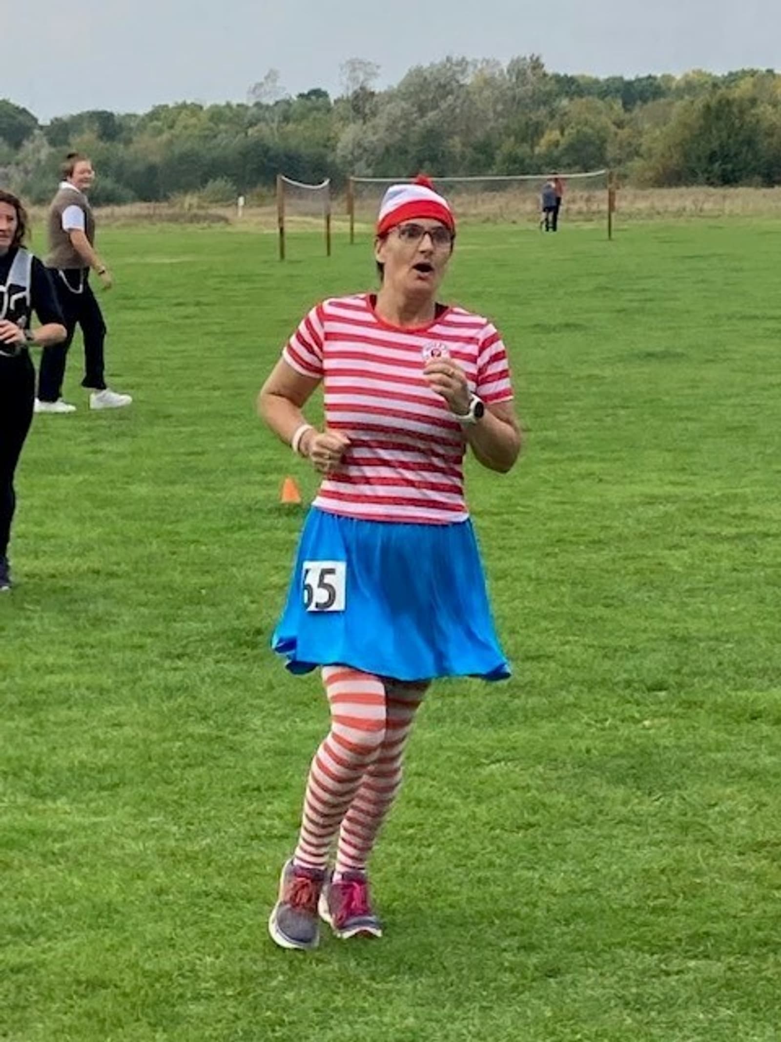 A runner, dressed in a red and white striped shirt, matching beanie, blue skirt, and red and white striped leggings, competes in a race on a grassy field. The runner wears a race bib with the number 55. Other participants and spectators can be seen in the background.