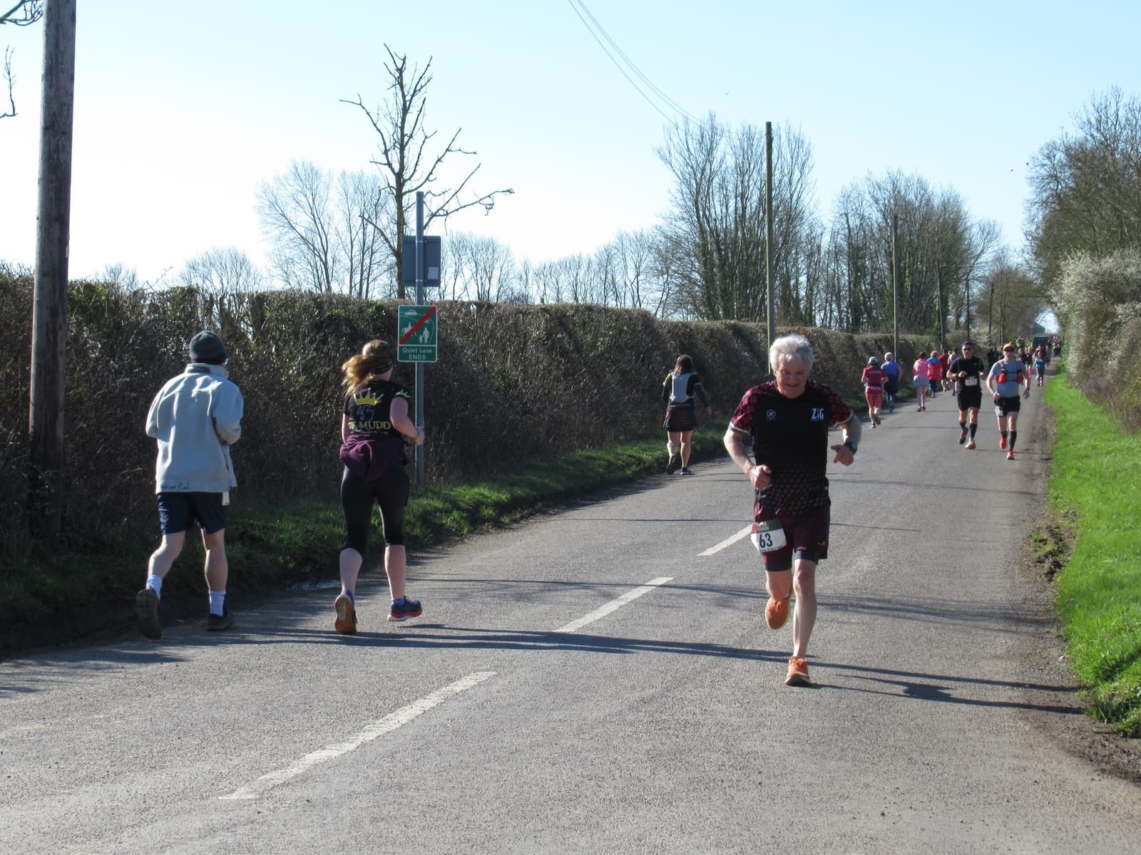 A group of runners participating in a road race during a sunny day. The lead runner, wearing a maroon outfit, runs on the right side of the road. Other runners follow behind on the left side, while bare trees and bushes line the road.