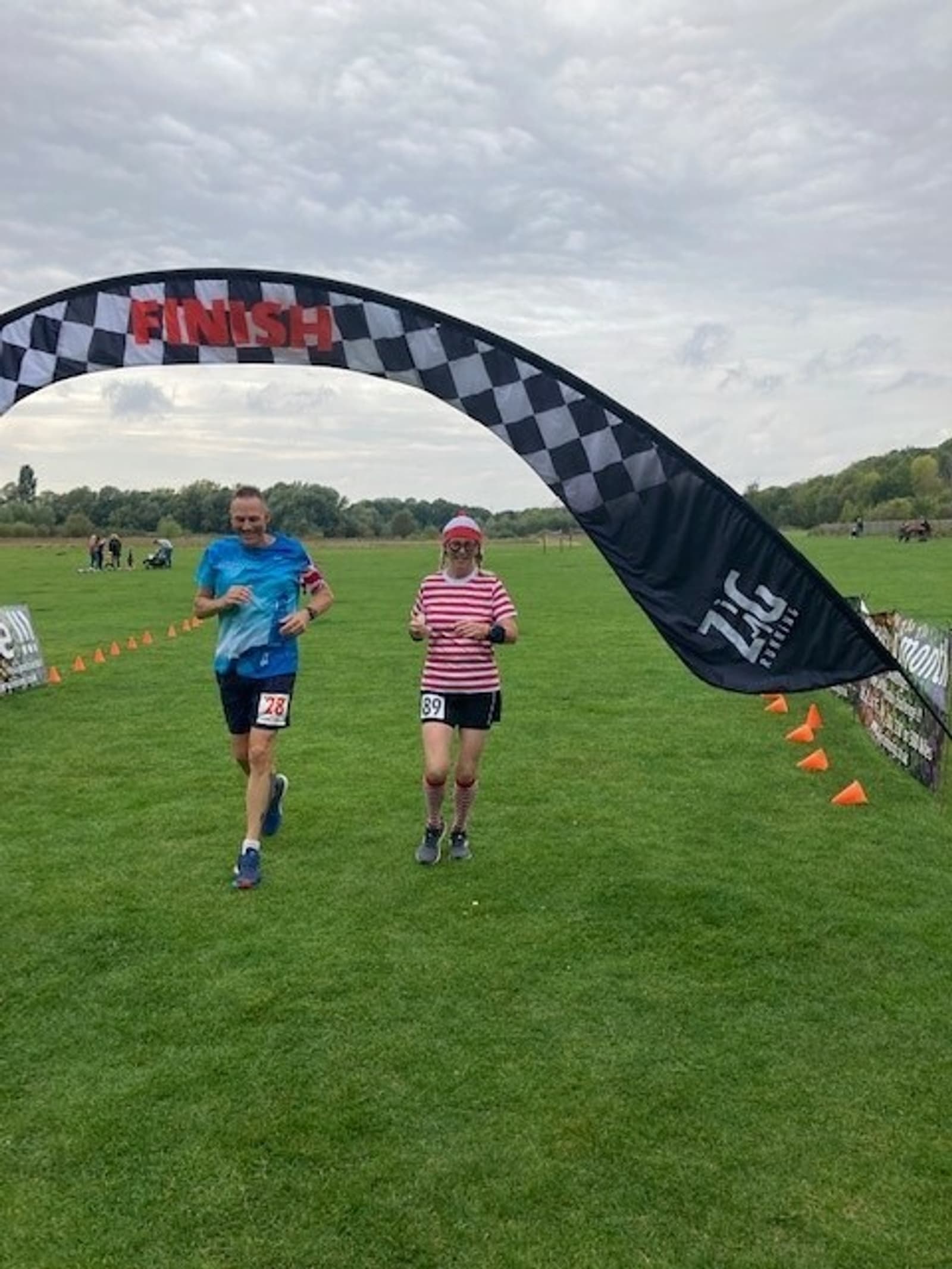 Two runners, one in a blue shirt and the other in a striped shirt and pink hat, cross a checkered finish line banner at a grassy event. Both are smiling, and orange cones and other participants are visible in the background on an overcast day.