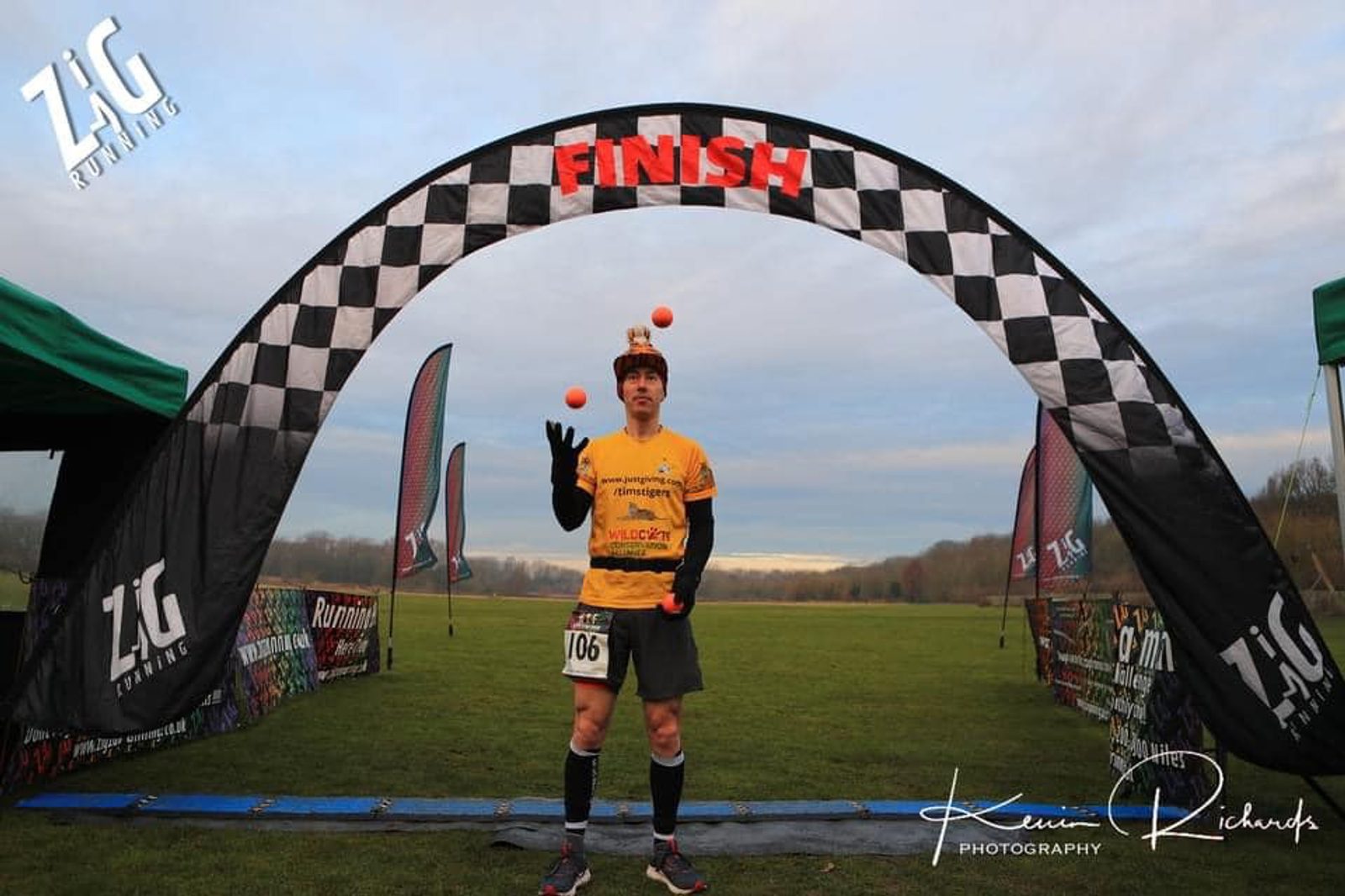 A man wearing an orange shirt, holding a trophy, is juggling three orange balls under a black and white checkered finish line archway at an outdoor running event. Banners and tents are set up on the grass around him.