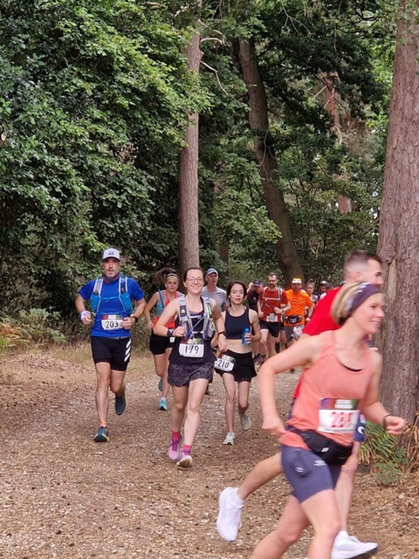A group of runners participating in a forest trail race. They are wearing race bibs and athletic gear as they run along a dirt path surrounded by tall trees and greenery. The runners appear focused and determined, with some smiling and interacting.