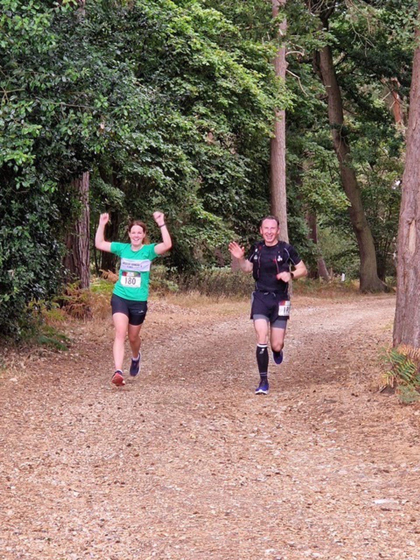 Two runners are on a forest trail, surrounded by tall trees and greenery. The runner on the left is wearing a green shirt and has both arms raised in victory. The runner on the right is in dark athletic wear, smiling and waving. The path is covered in dry leaves.