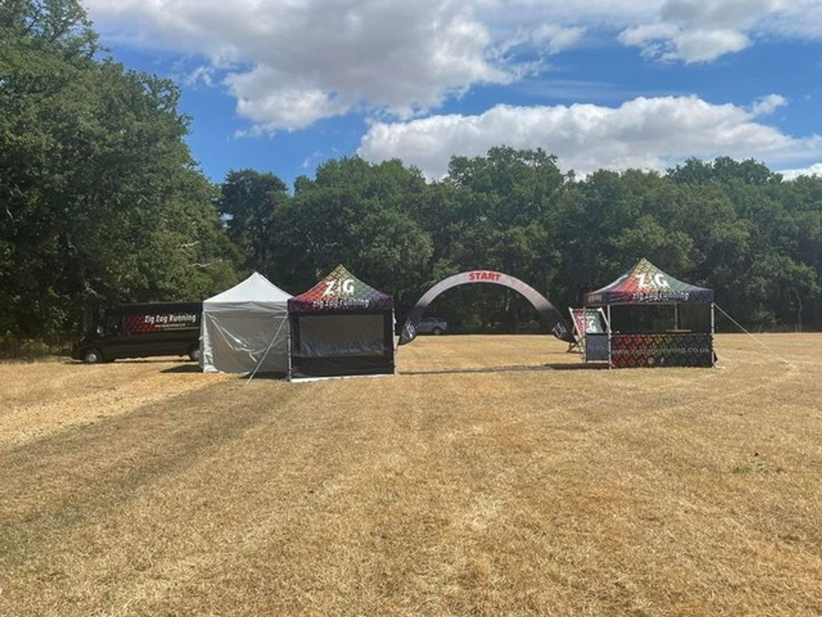 Outdoor event setup in a grassy field with two branded tents labeled "Zig Zag." An adjacent van also with "Zig Zag" branding is situated on the left. A large archway marked "Start" is in the center, and trees and a partly cloudy sky form the backdrop.