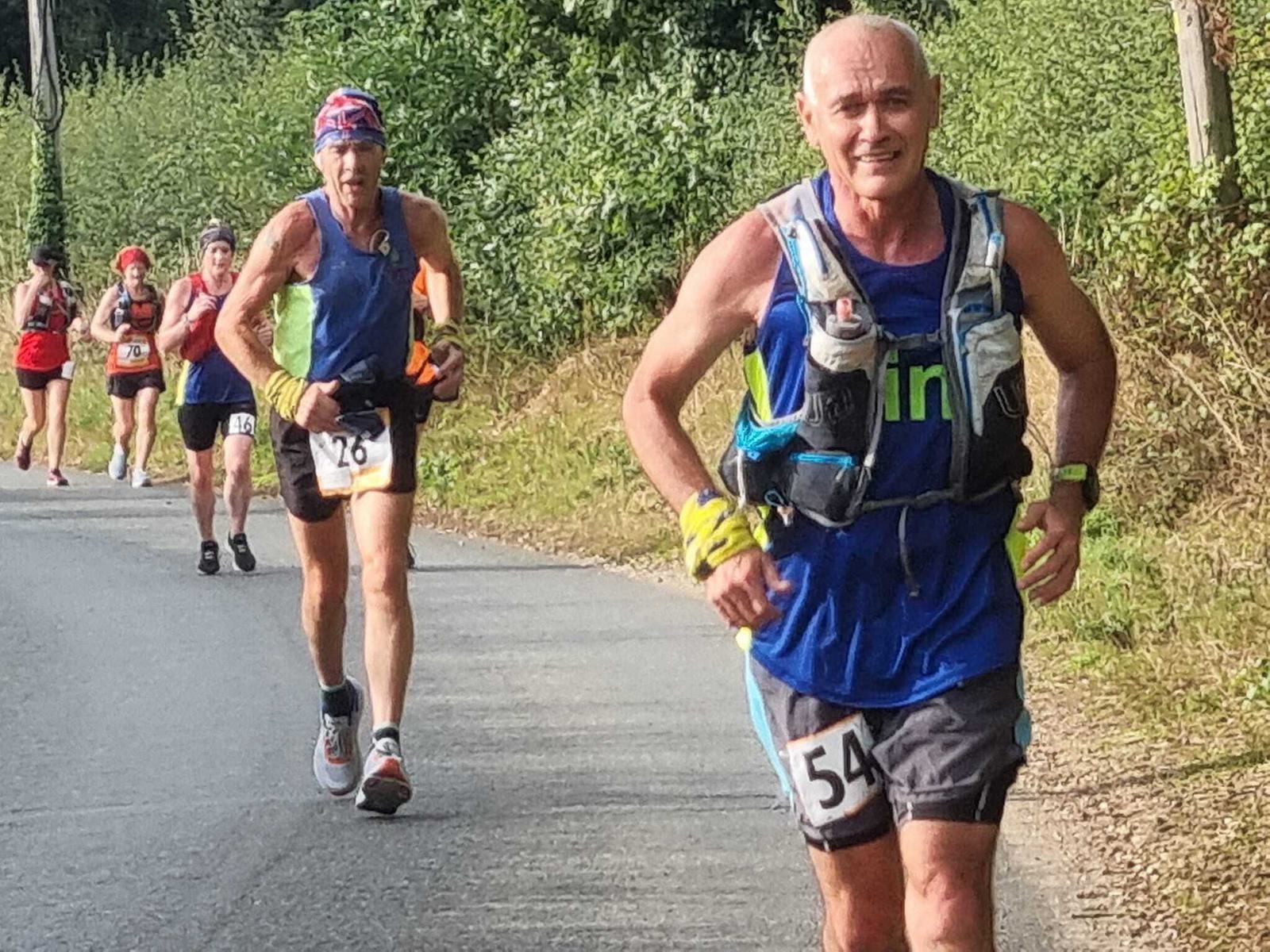 A group of marathon runners, some wearing hydration packs and numbered bibs, race on a paved road surrounded by lush greenery. The runner in front, wearing bib number 54 and a blue tank top, appears focused, with other runners close behind.