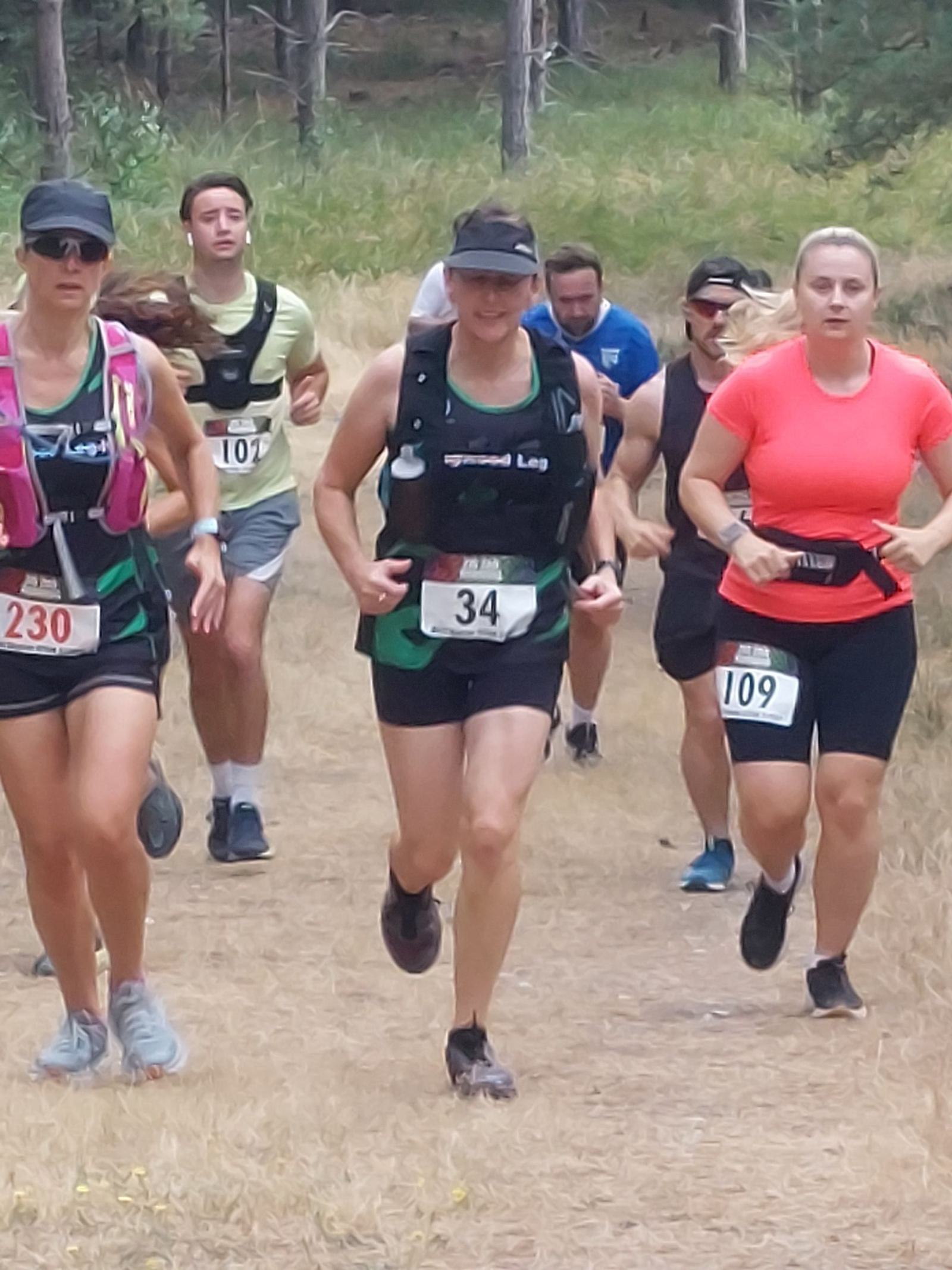 A group of runners participating in a trail race through a forest. Several runners are wearing numbered bibs and hydration packs. The path is surrounded by trees and grass, and the atmosphere appears focused and energetic.