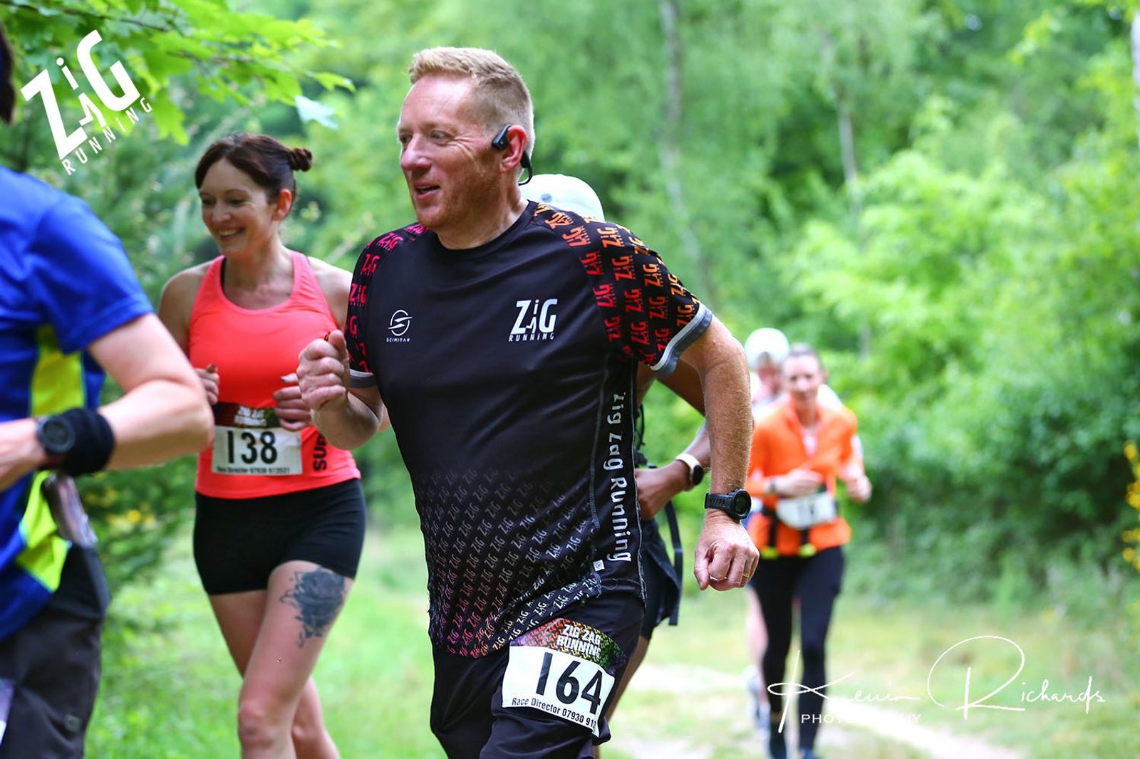 A group of runners jogs on a forest trail during a race. The lead runner wears a black and red Zig Running shirt and has the number 164 on his race bib. Other participants in colorful athletic wear can be seen in the background, surrounded by lush greenery.