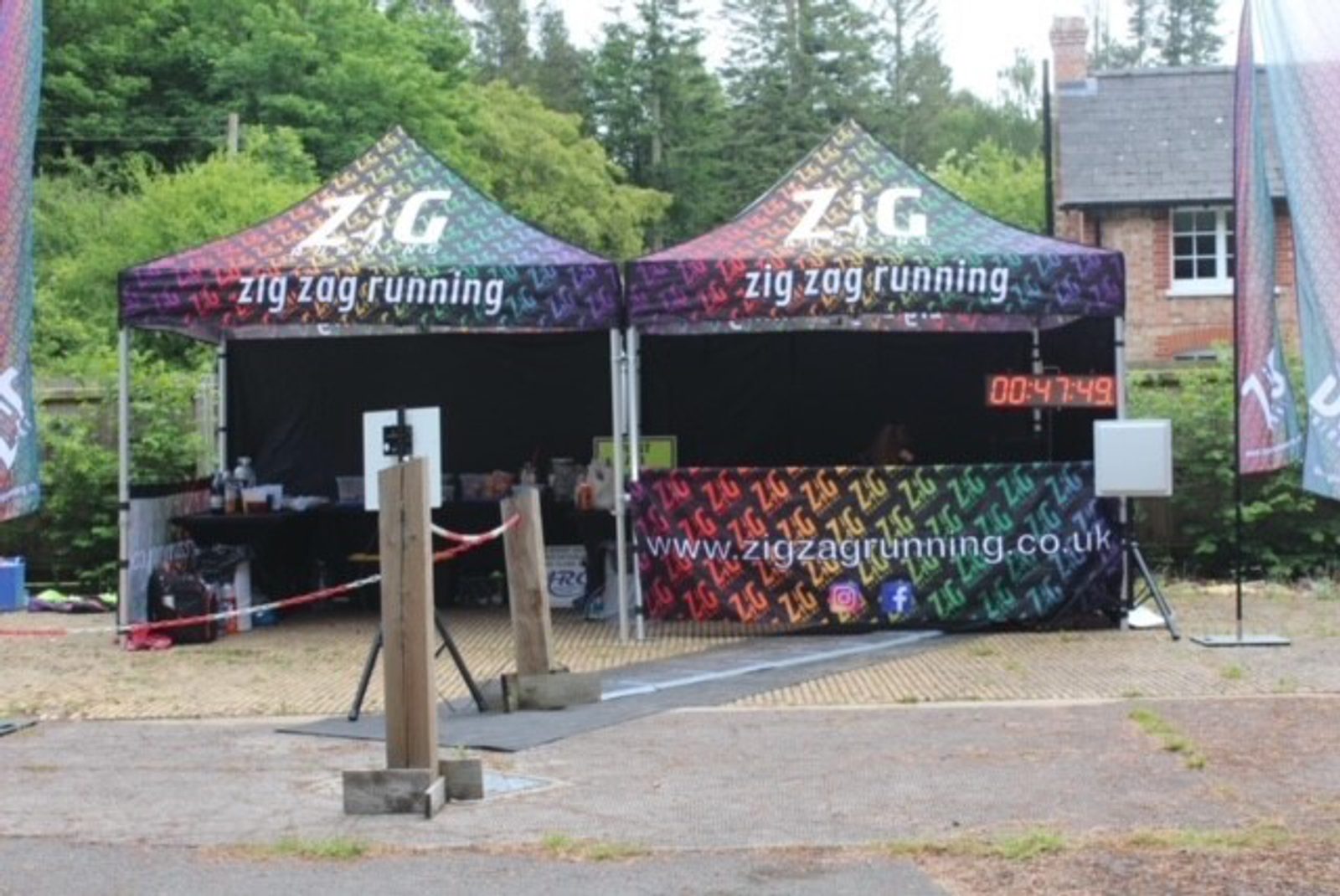 Two black tents with the text "Zig Zag Running" and "www.zigzagrunning.co.uk" printed on them are set up at an outdoor event. There is a digital timer displaying "00:47:49." The tents are surrounded by trees and a building in the background.