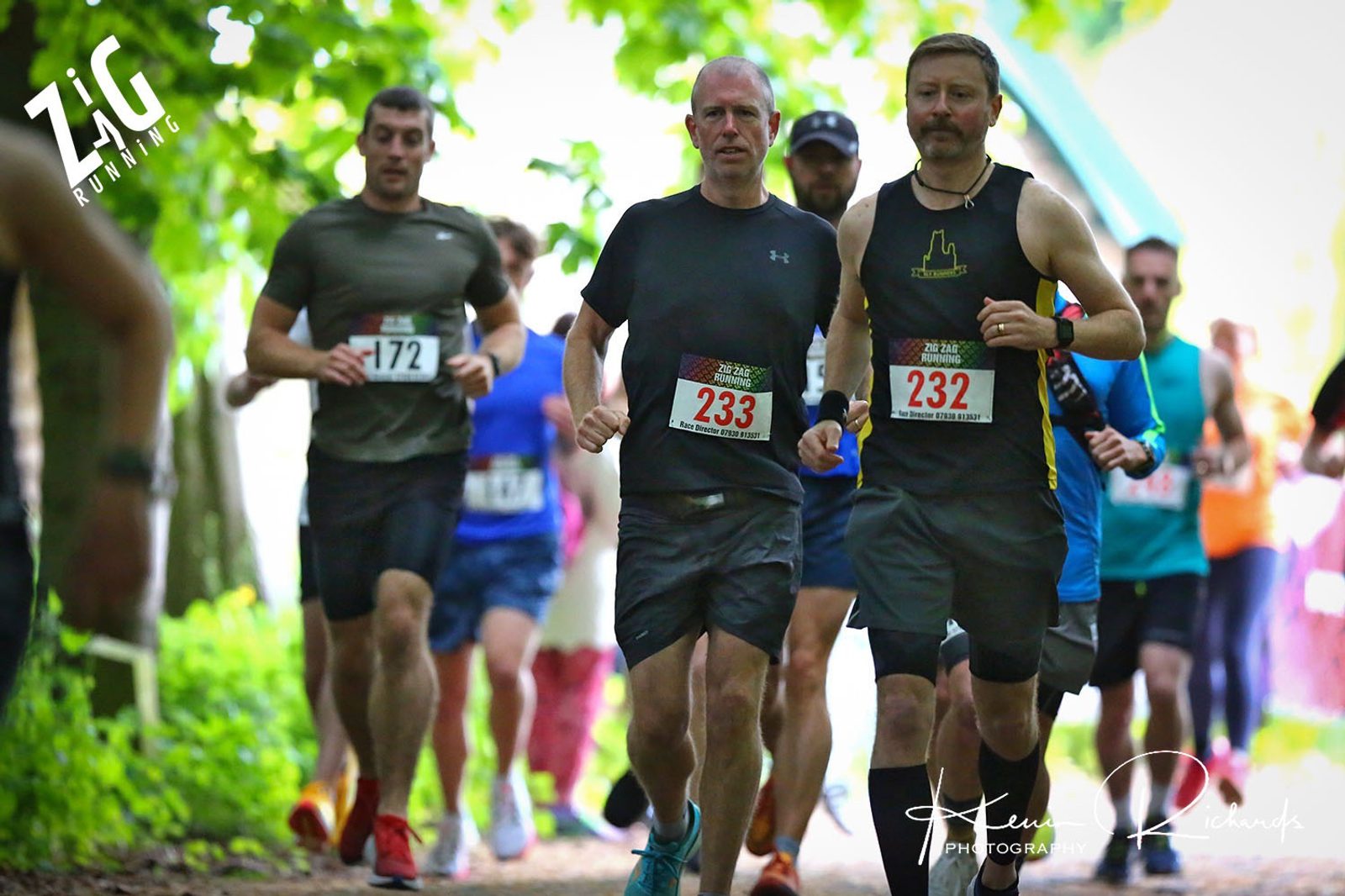 Runners participate in an outdoor race, moving along a wooded trail. Two men in the foreground wear bib numbers 233 and 232, respectively. Other runners can be seen in the background under dappled sunlight. The logo "Zigzag Running" is visible in the top left corner.