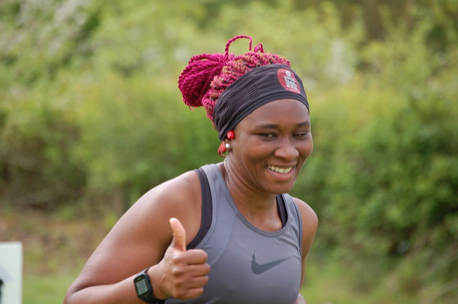 A woman with braided hair, a black headband, and a grey sleeveless athletic top is smiling and giving a thumbs-up gesture while jogging outdoors. She has a fitness tracker on her wrist, and the background is blurred greenery.