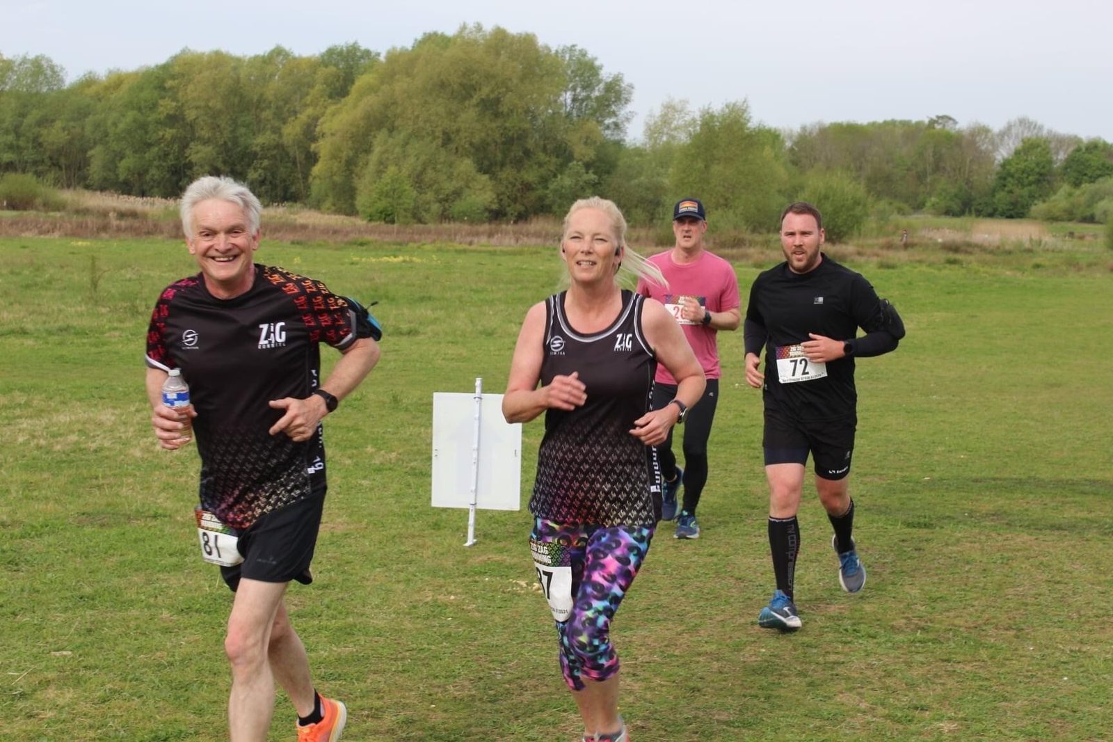A group of four people run on a grassy path during a race. Two people in the front are smiling, wearing matching black athletic outfits. A man in a pink shirt and another person in black follow closely behind. Trees and bushes are visible in the background.
