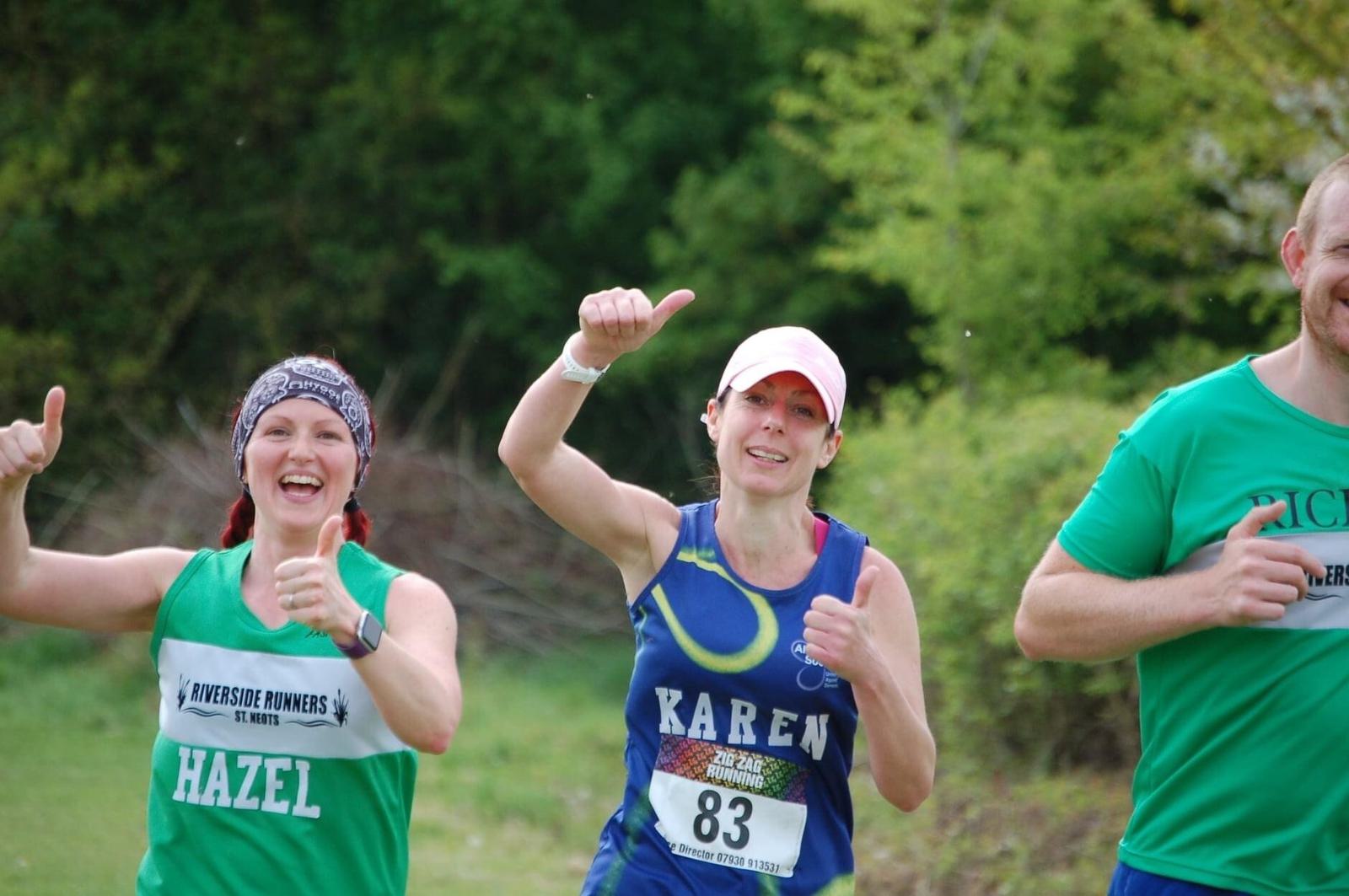 Three runners smile and give thumbs up during a race. The woman on the left, wearing a headband and "Hazel" bib, and the woman in the middle, wearing a pink cap and "Karen" bib, both look energized. A man in a green shirt is on the right, partially cropped out.
