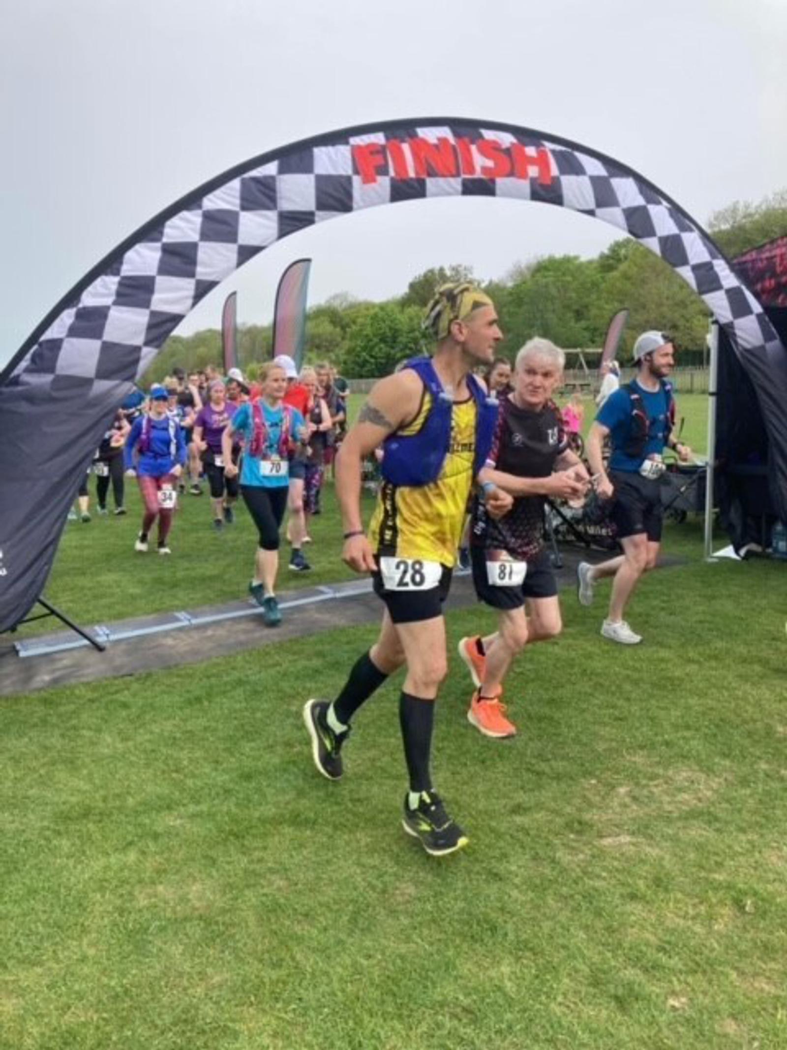 Runners are crossing a black and white checkered finish line arch. The runner in the foreground wears a yellow and blue outfit with bib number 28. Beside him is a runner with bib number 81 in a black outfit. Other runners and spectators are visible in the background.