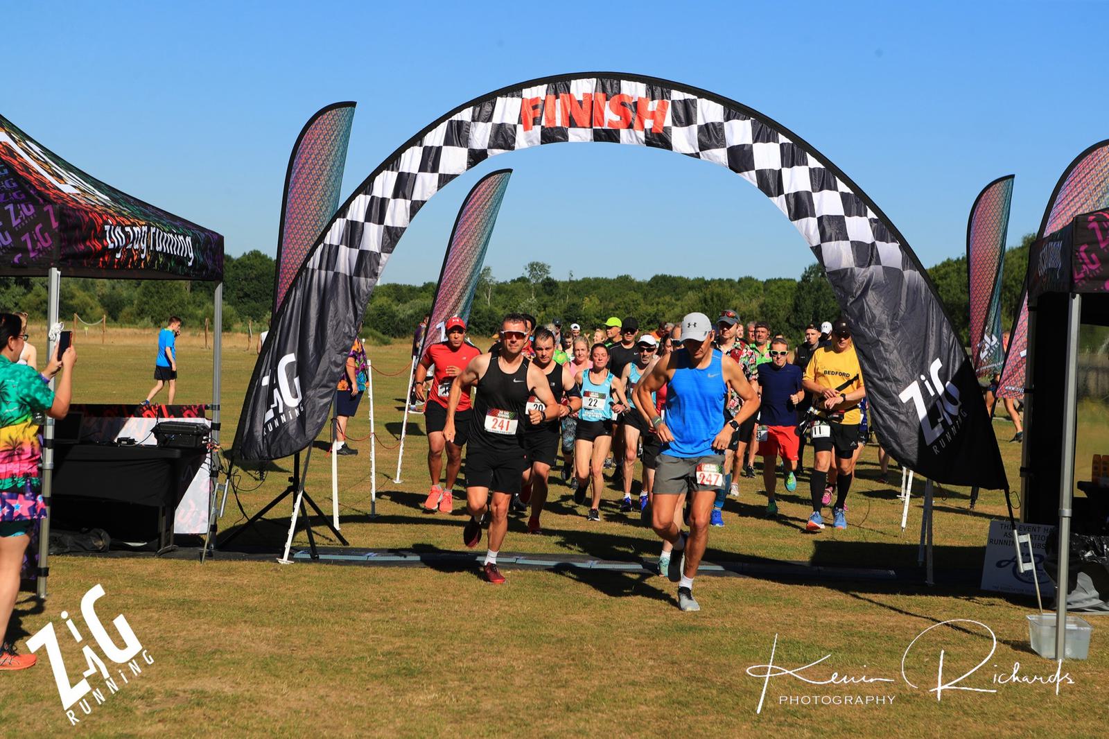 Runners cross the starting line beneath a checkered arch at an outdoor race event. Participants in various running attire are seen on a grassy field, surrounded by tents and banners with the event's branding. The background shows a clear blue sky and trees.