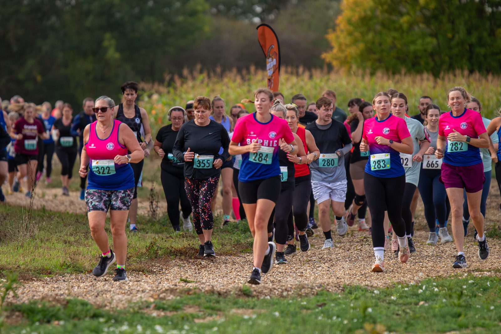 A group of runners, many wearing numbered bibs and pink shirts, participate in an outdoor race on a gravel path surrounded by greenery. The atmosphere appears energetic and focused.