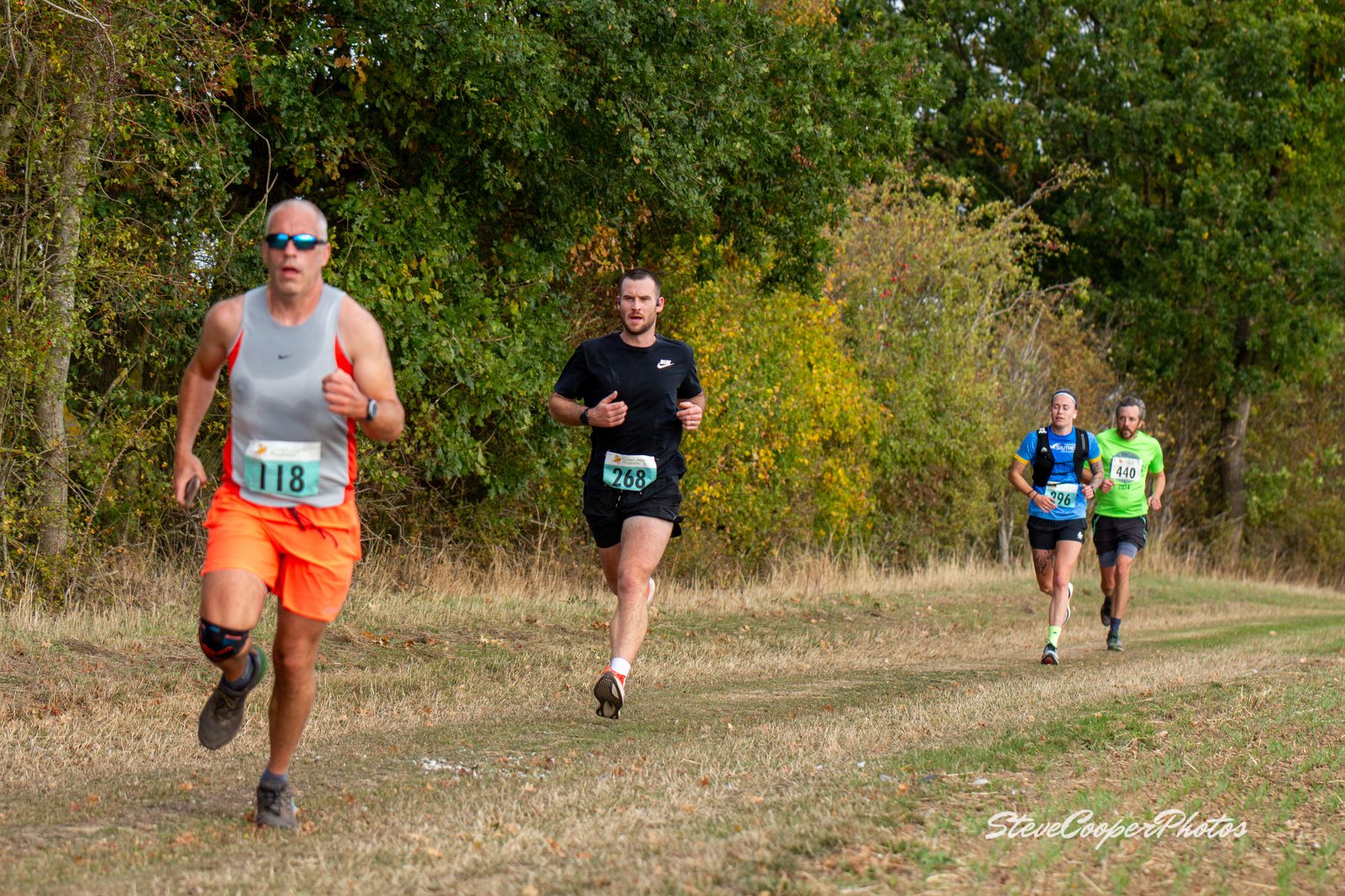 Four runners, each wearing numbered bibs, compete on a grassy trail beside trees. The runner in front wears sunglasses and an orange knee brace. The other three follow, spaced apart, with trees and autumn foliage in the background.
