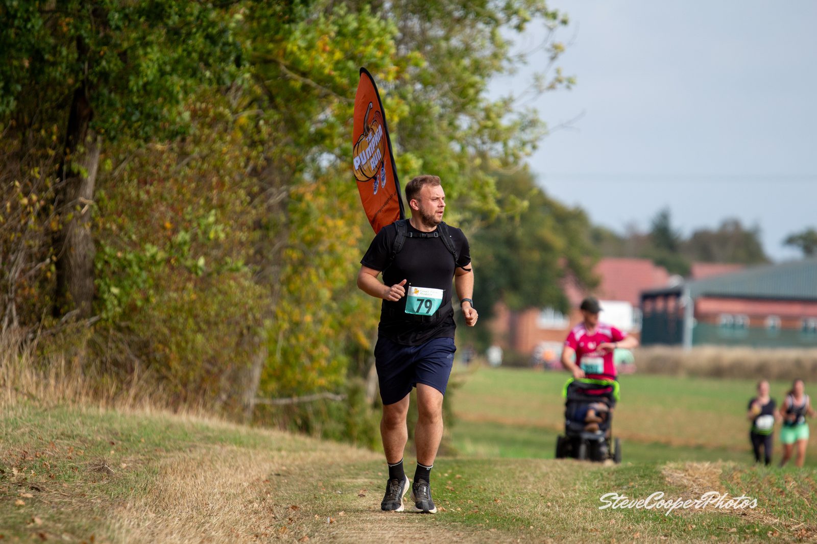 A man wearing a race bib number 79 runs on a grassy trail near trees. He carries an orange flag and looks focused. In the background, people and a stroller can be seen on the path.