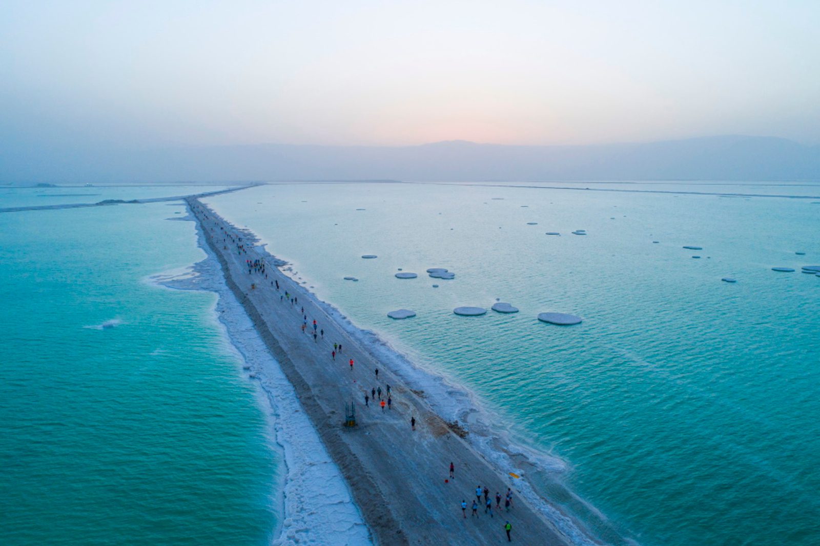 Aerial view of people walking along a narrow, salt-covered pathway that divides the turquoise waters of the Dead Sea. Circular salt formations are scattered across the water, and the hazy sky suggests either dawn or dusk.