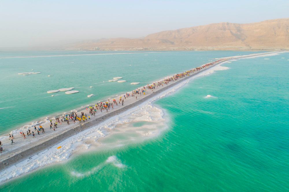 Aerial view of a long narrow strip of land surrounded by turquoise water, with a large group of people walking along it. The surrounding landscape includes salt formations and distant mountains under a clear sky.