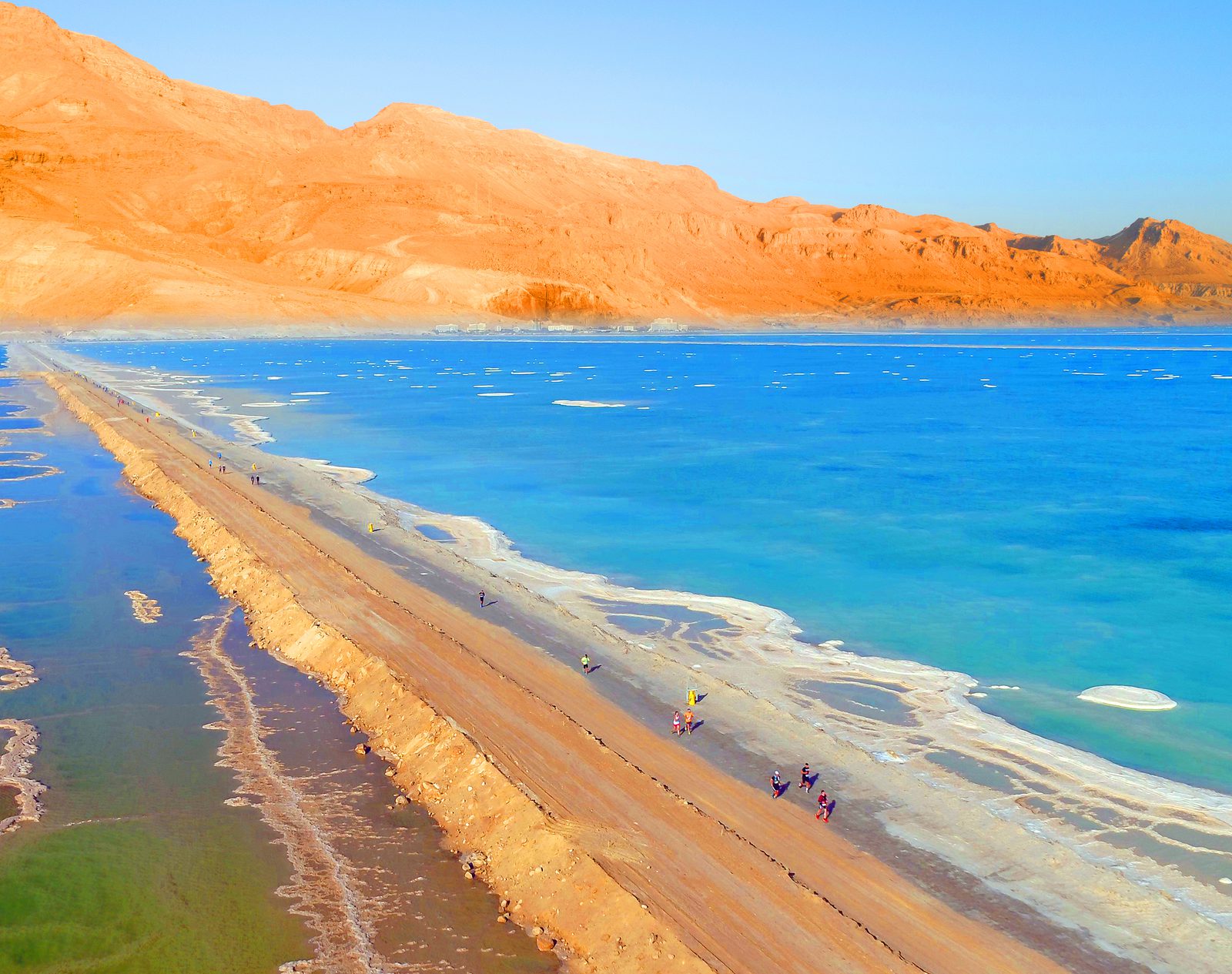 Aerial view of a narrow spit of land with people walking on it, surrounded by vibrant blue water on one side and still water on the other, with a backdrop of striking orange desert mountains under a clear blue sky.