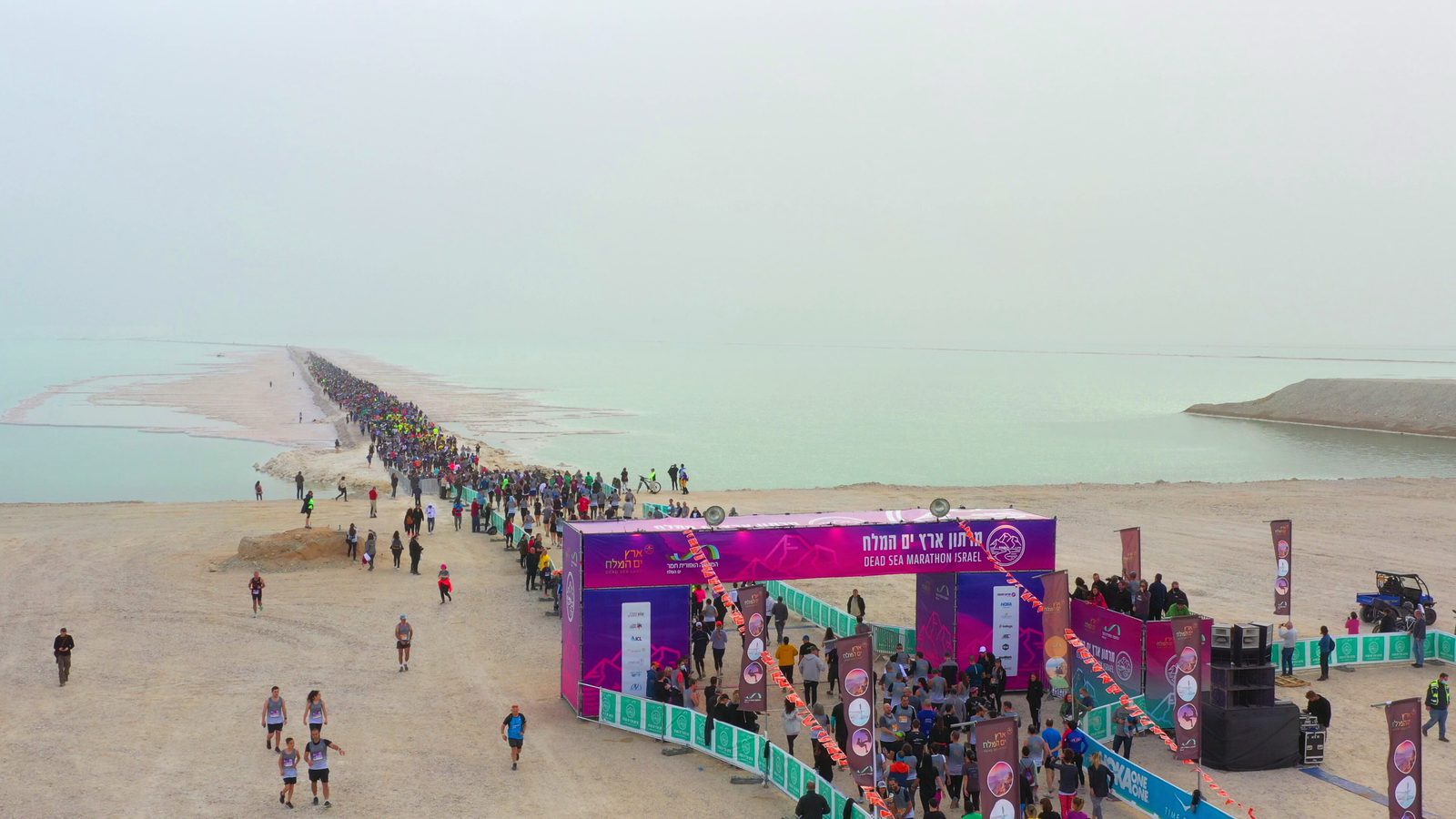 A large crowd of runners is gathered near the starting line of a marathon event on a scenic beach. The course stretches into the distance along the shoreline. Colorful banners and flags mark the starting area, and the ocean is visible in the background under an overcast sky.