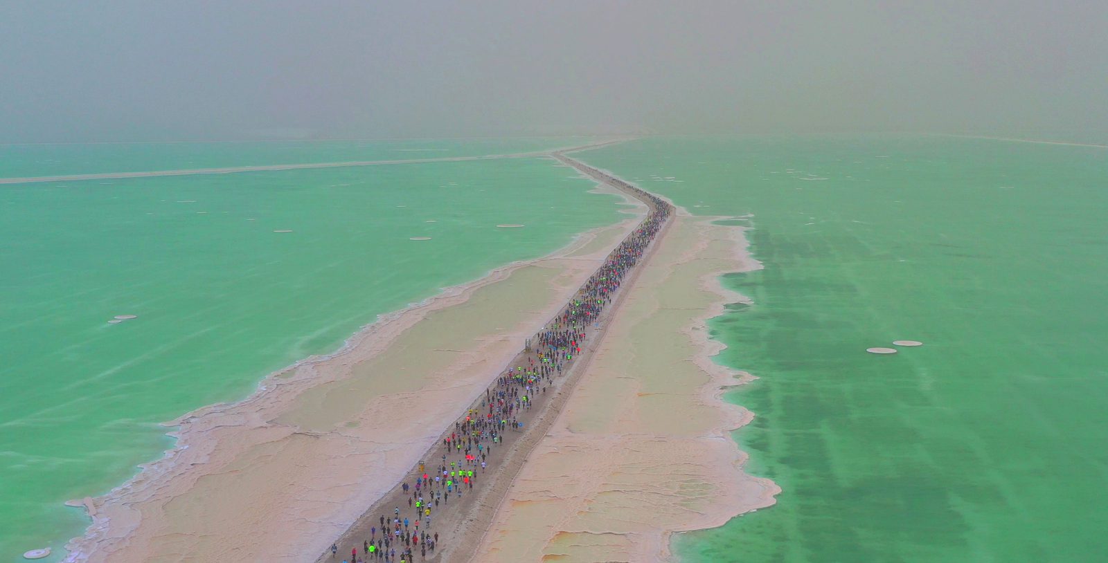 Aerial view of a long crowd of people walking along a narrow, sandy path surrounded by greenish-blue water. The path stretches into the distance, fading into the misty horizon. The scene has an ethereal quality.