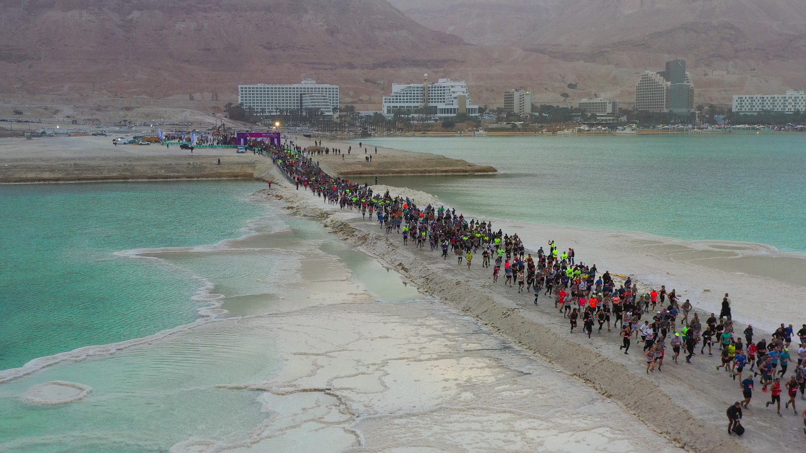 A large group of runners participate in a marathon on a narrow strip of land between emerald green waters. In the background, a desert landscape and several modern buildings are visible. The sky appears overcast, casting a muted light over the scene.