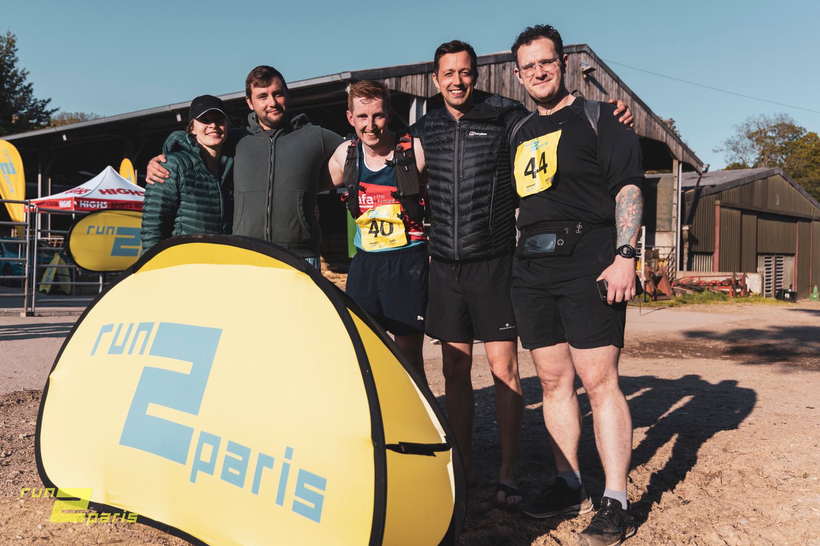 Five people are standing together and smiling in front of a yellow "Run 2 Paris" banner. Three of them are wearing race numbers and athletic clothing, while two are in casual attire. The background shows a rustic building and race-related paraphernalia.