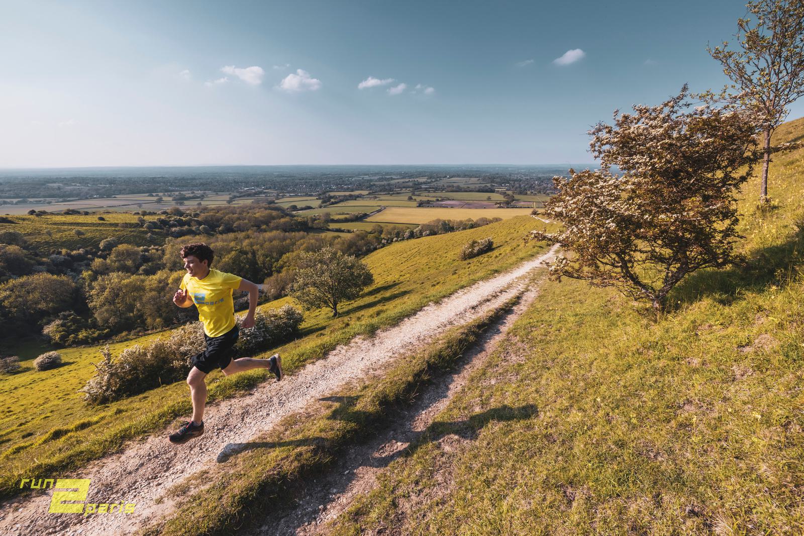 A man in a yellow shirt runs uphill on a dirt trail through a grassy, open landscape under a clear blue sky. A tree is on the right, and a patchwork of fields stretches into the distance. The words "run dream create" are seen in yellow on the bottom left.