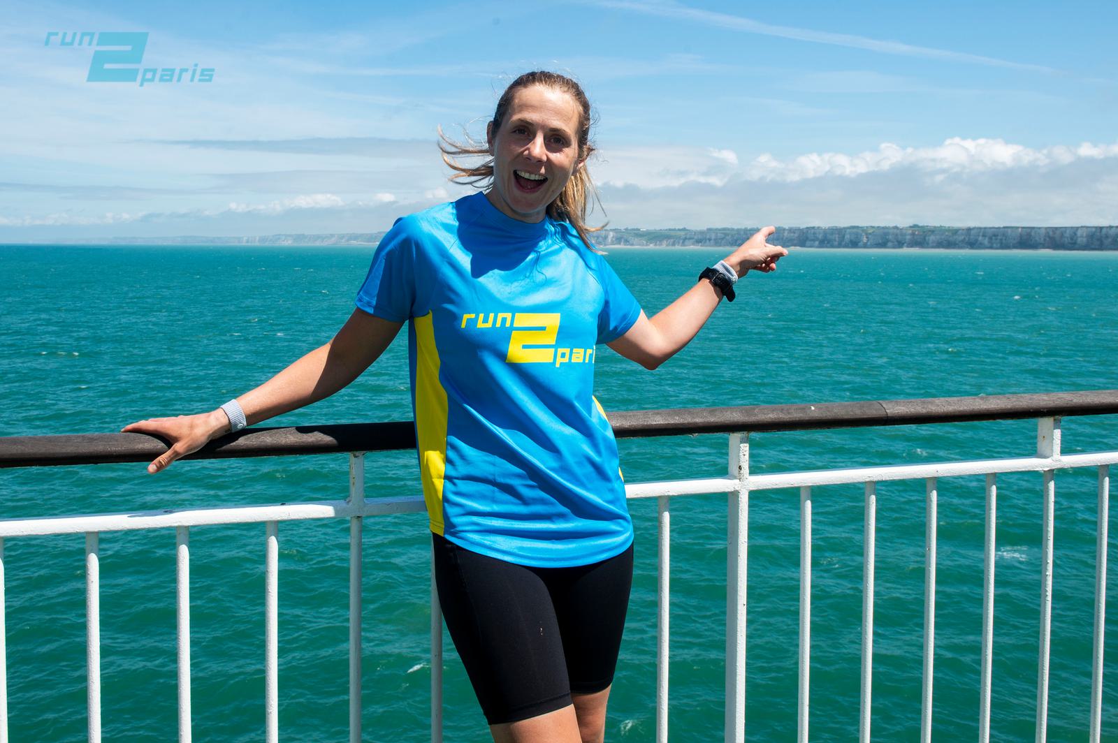 A person wearing a blue "run2paris" shirt and black shorts is smiling and pointing towards the sea. They are standing on a ferry's railing with an ocean and distant coastline in the background on a sunny day.