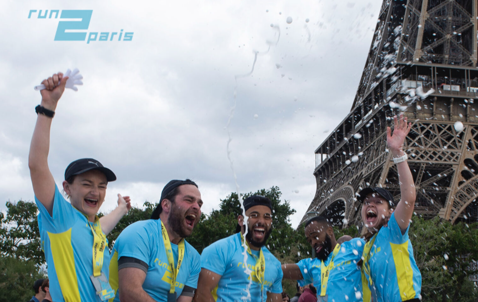 A group of five people in matching blue shirts celebrate enthusiastically in front of the Eiffel Tower while participating in the "Run 2 Paris" event. One person is holding a water bottle that is spraying water, adding to the festive atmosphere.