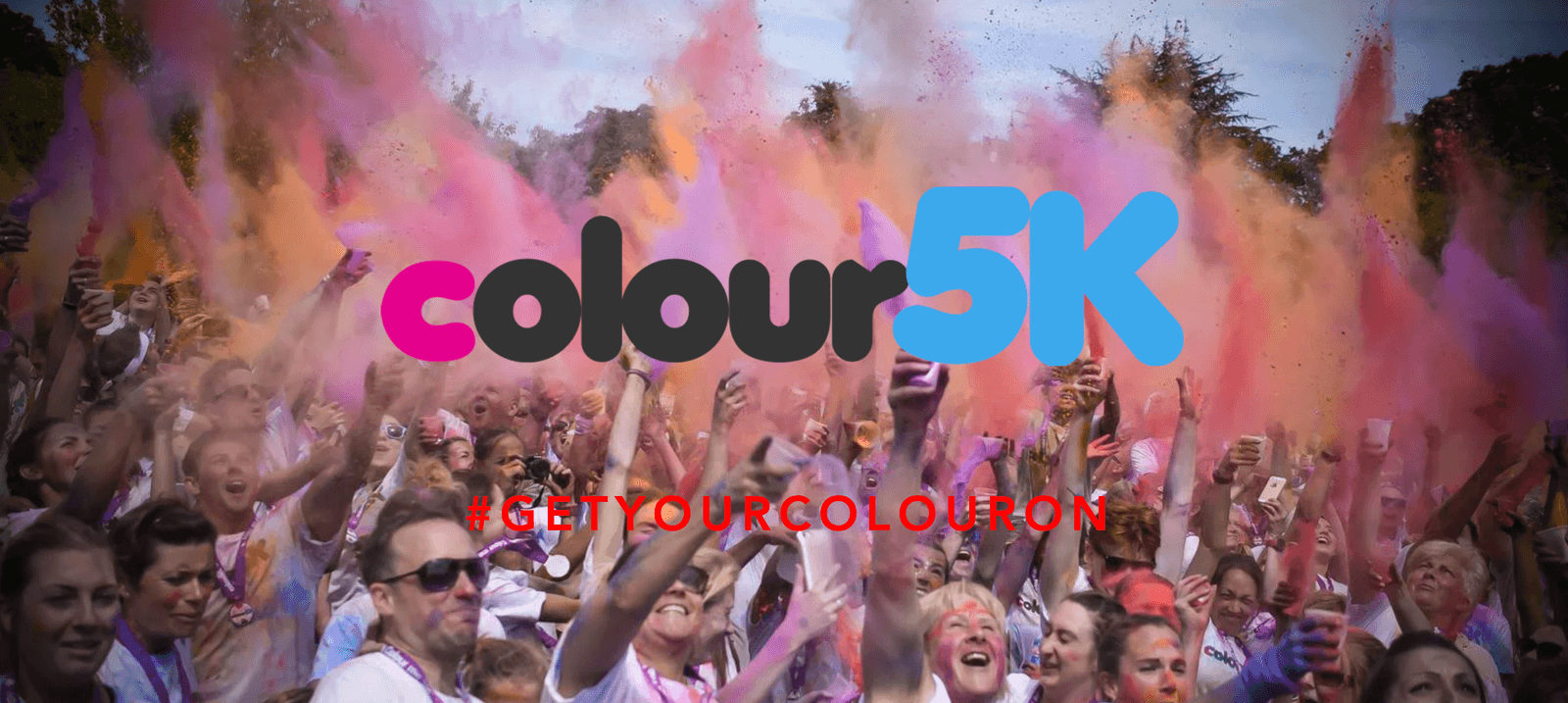 A crowd of enthusiastic participants in white t-shirts throw vibrant colored powders into the air at a Color Run event. The words "colour 5K" appear prominently in the center with the hashtag #GETYOURCOLOURON below. The scene is filled with joy and vibrant colors.