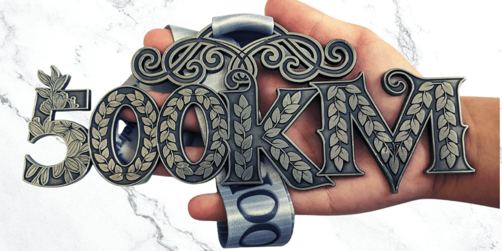 A hand holds a silver medal with intricate floral designs and the text "500KM" prominently displayed on it. The background is a white marble surface.