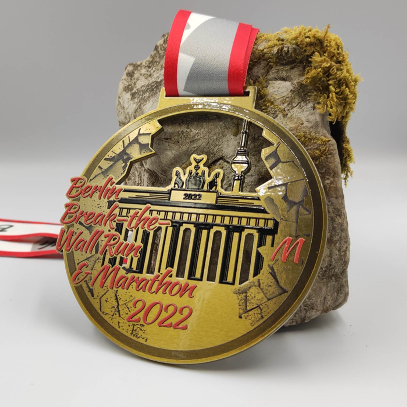 A gold medal with a red and white ribbon features an embossed design of the Brandenburg Gate and Berlin TV tower, with "Berlin Break the Wall Run & Marathon 2022" written in red text. The medal is placed against a rock adorned with small patches of moss.