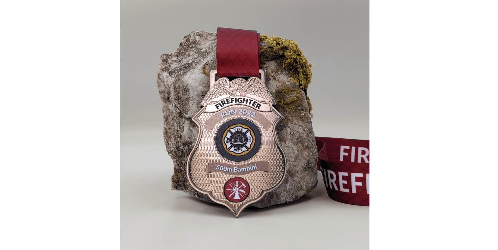 A firefighter-themed medal with a red ribbon is resting against a rock. The medal reads "FIREFIGHTER RUN 2022" at the top, with "500m Bambini" below a central firefighter emblem. A rolled red strap, partially visible, lies next to the rock.