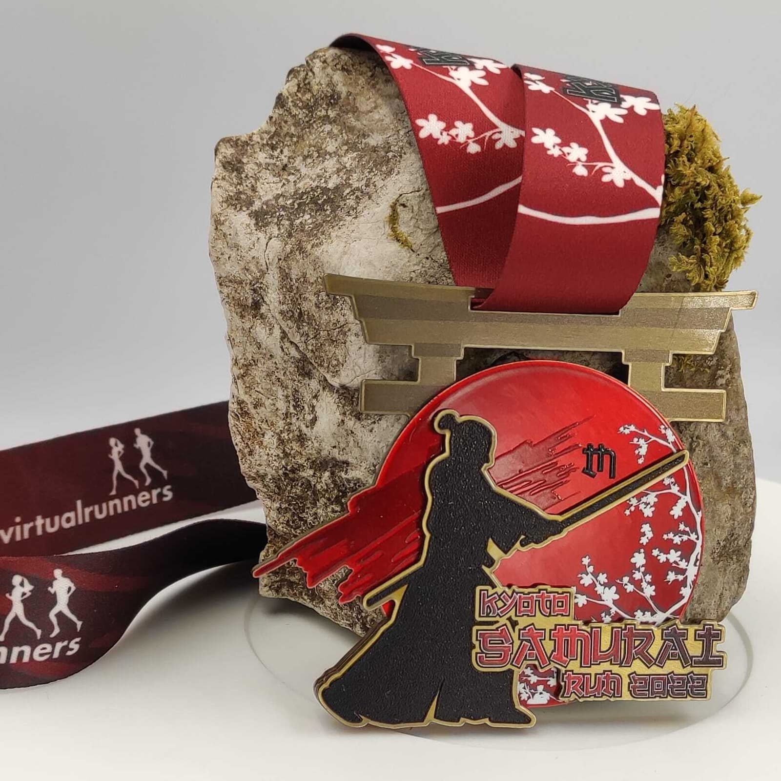 A medal with a red and white ribbon draped over a rock. The medal features a torii gate and a samurai silhouette with a sword against a red sun background. The ribbon has white runner icons and the text "virtualrunners." The medal reads "Tokyo Samurai Run 2023.