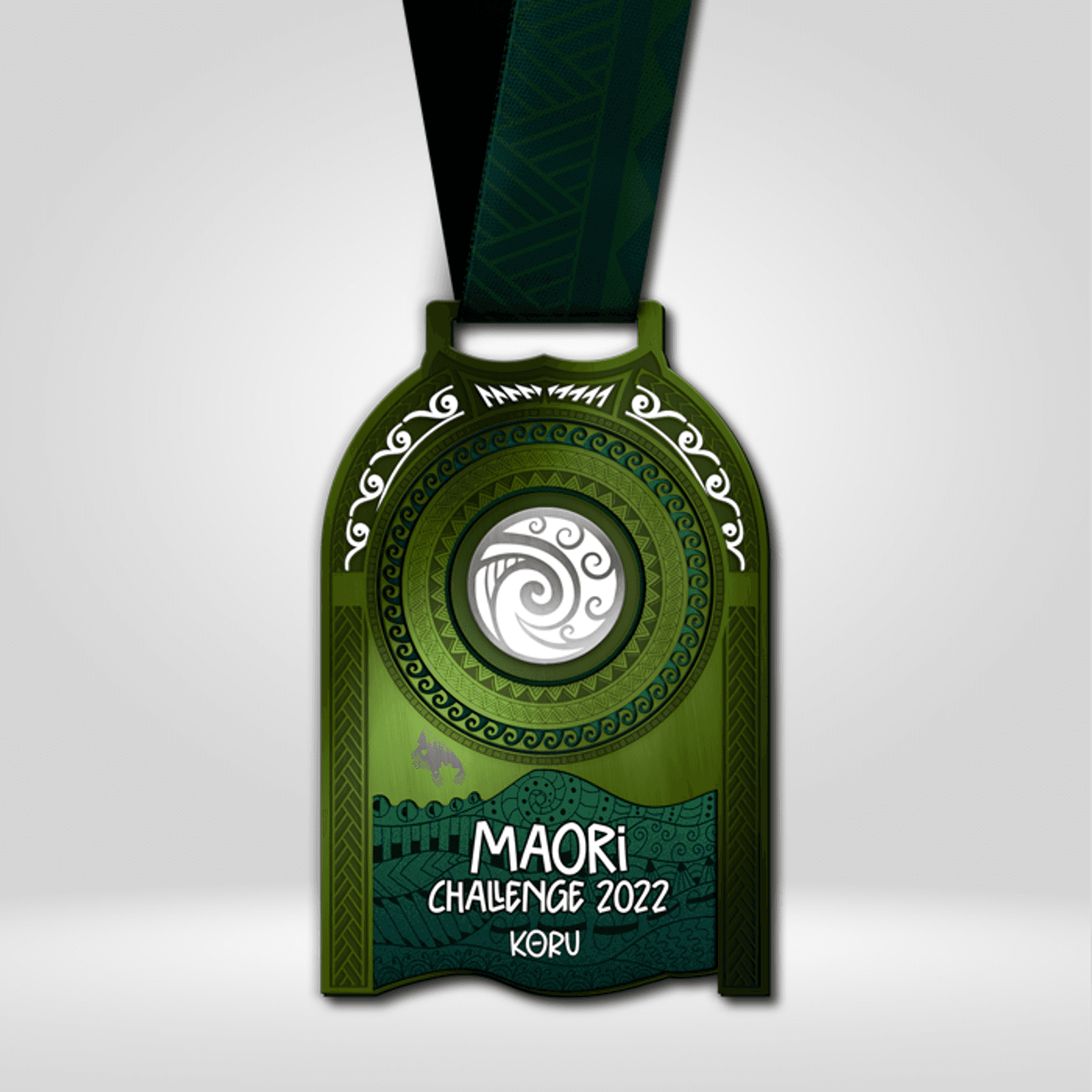 A green medal with intricate MΔori designs hangs from a dark green ribbon. The central motif is a white koru spiral framed by patterns. Below, white text reads "MΔori Challenge 2022 Koru" against a textured green background with additional tribal art.